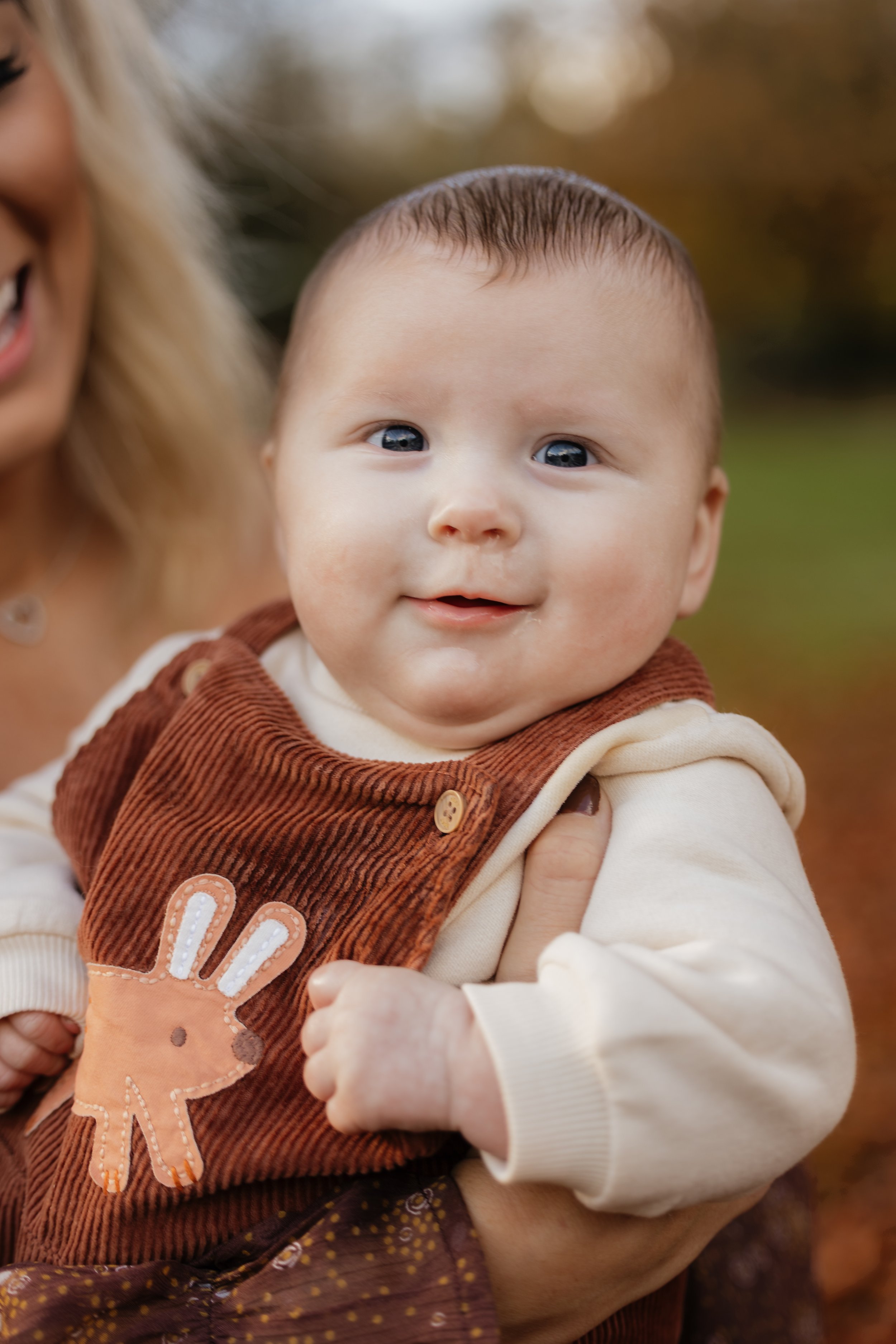A smiling baby with blue eyes in a brown and cream outfit being held outdoors during fall.