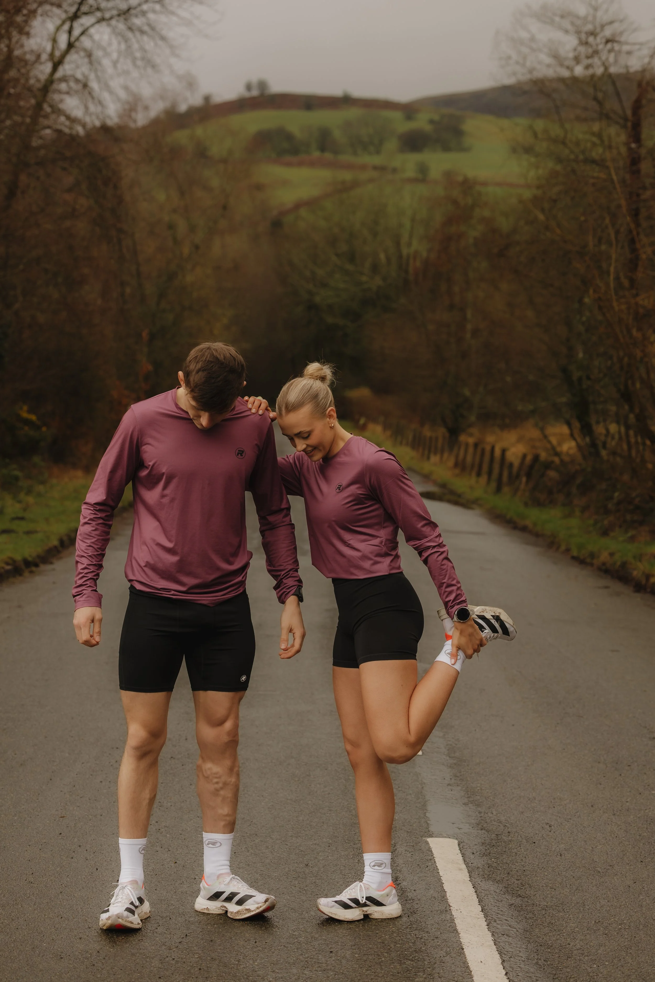 A man and woman in athletic clothing stretch on an empty rural road surrounded by trees and hills.