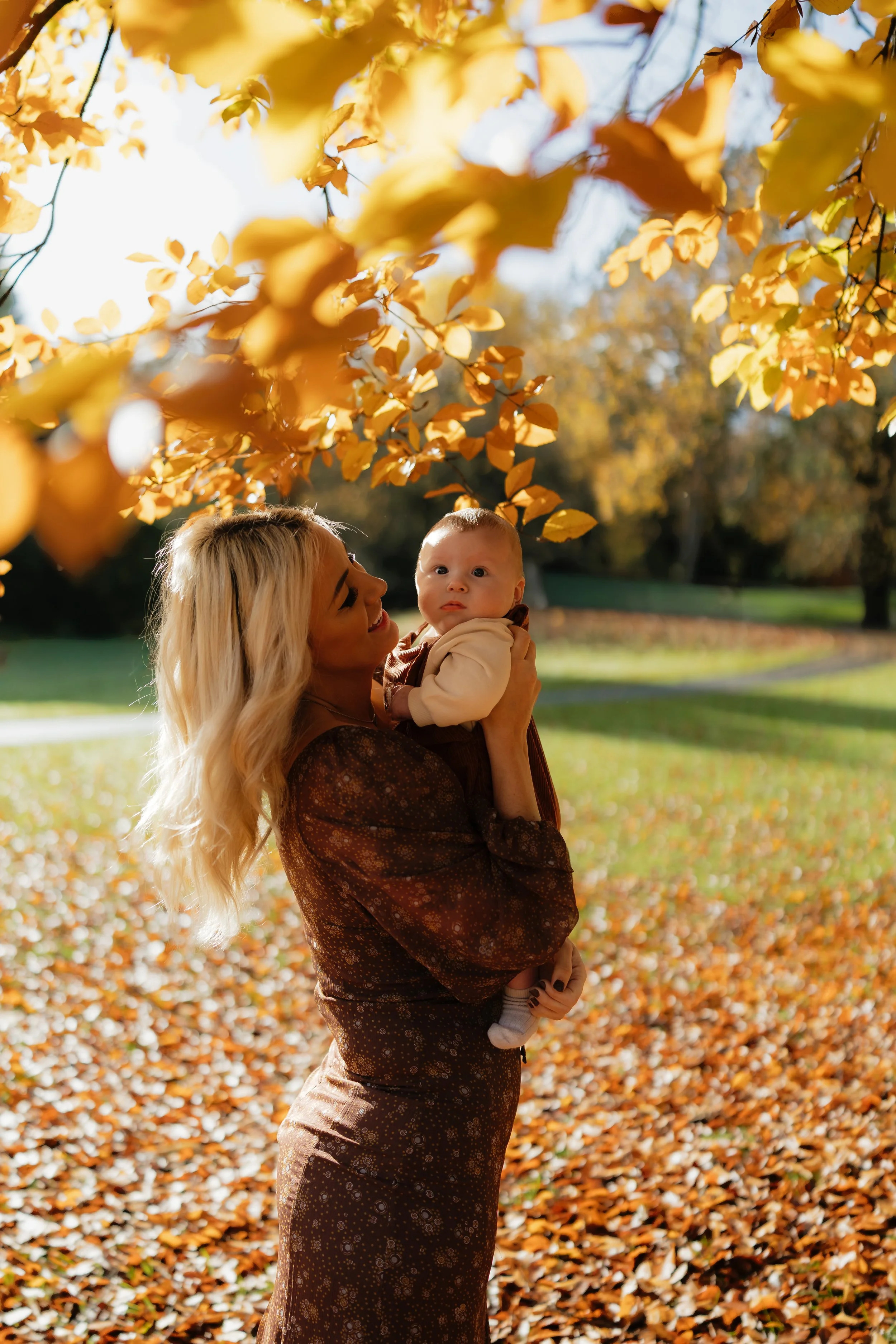 A woman holding a baby outdoors during autumn, with fallen leaves on the ground and yellow leaves on trees.