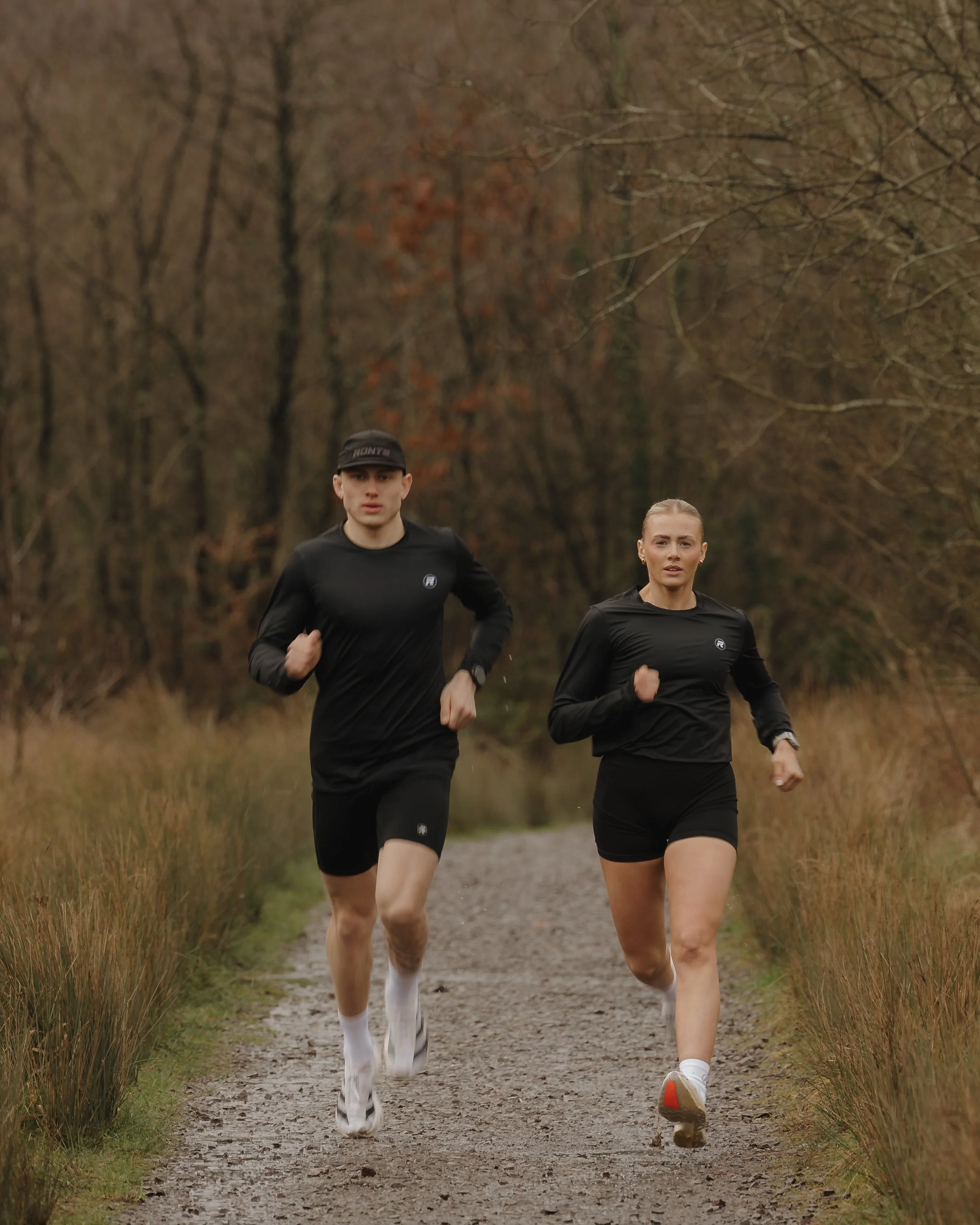 Two people running on a dirt trail in a wooded area during fall.