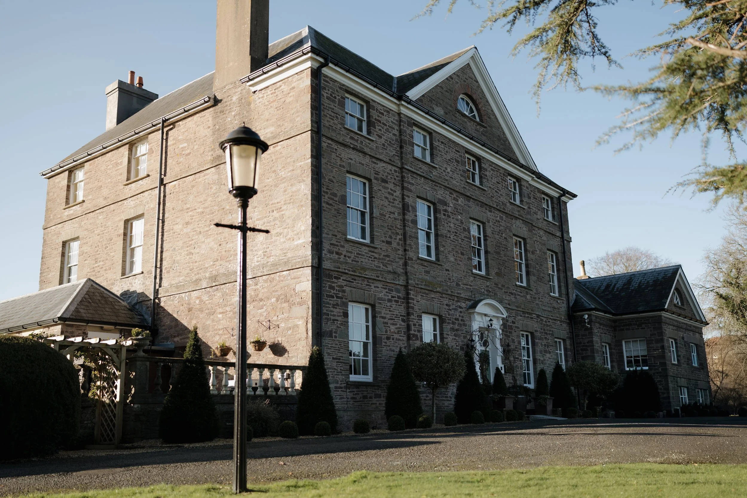 Large historic brick house with multiple windows, a white entrance door, and surrounded by landscaped bushes and trees. A black lamppost is in the foreground, and a clear blue sky is above.