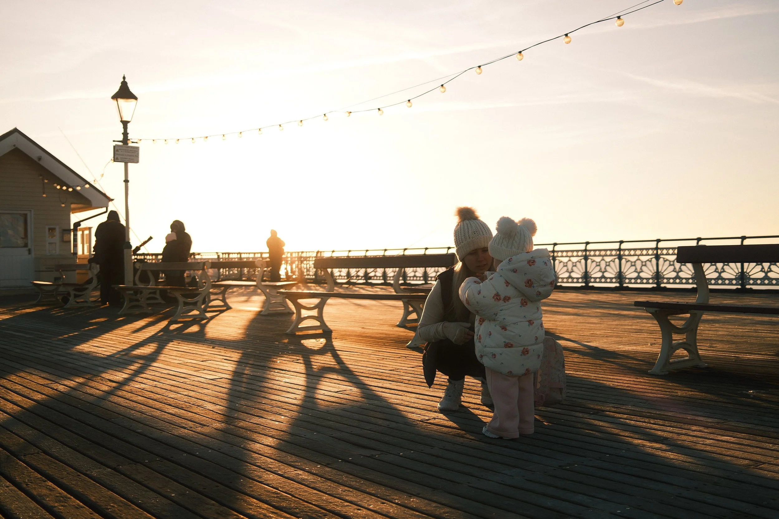 A woman and a young child on a wooden pier during sunset, with string lights overhead, benches, a small building, and a railing in the background.