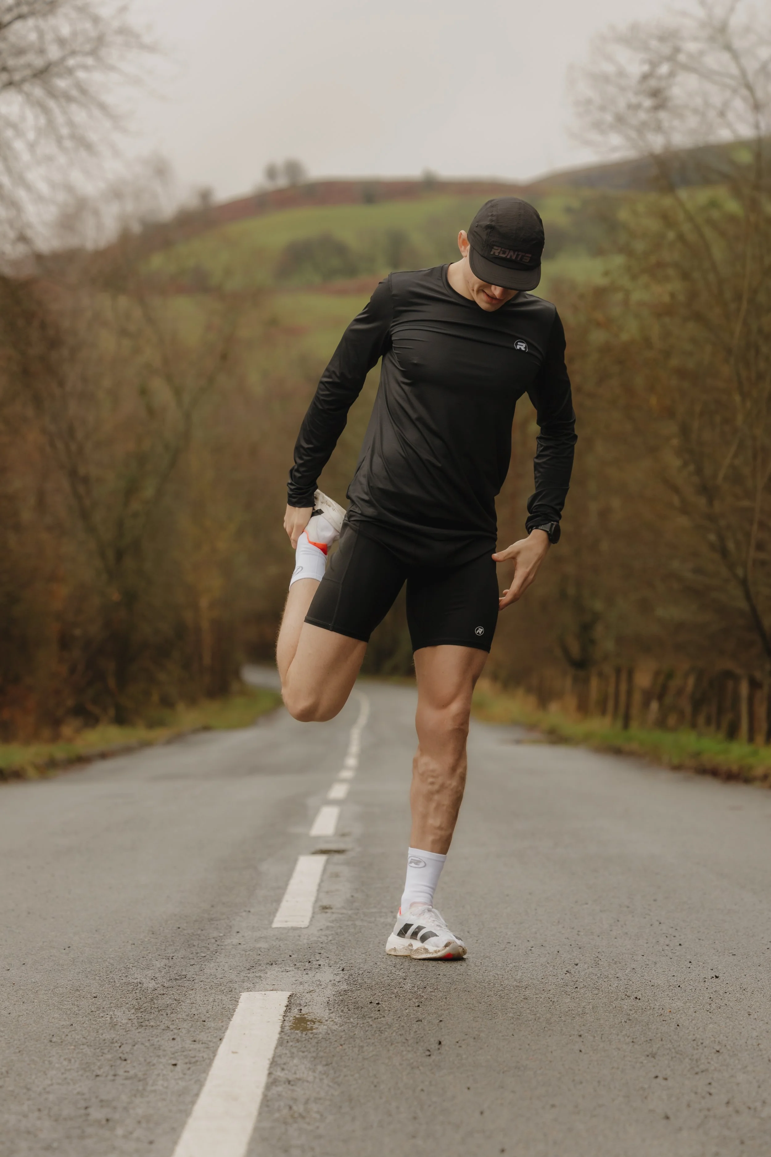 A man stretching his leg on an empty country road surrounded by trees and rolling hills.