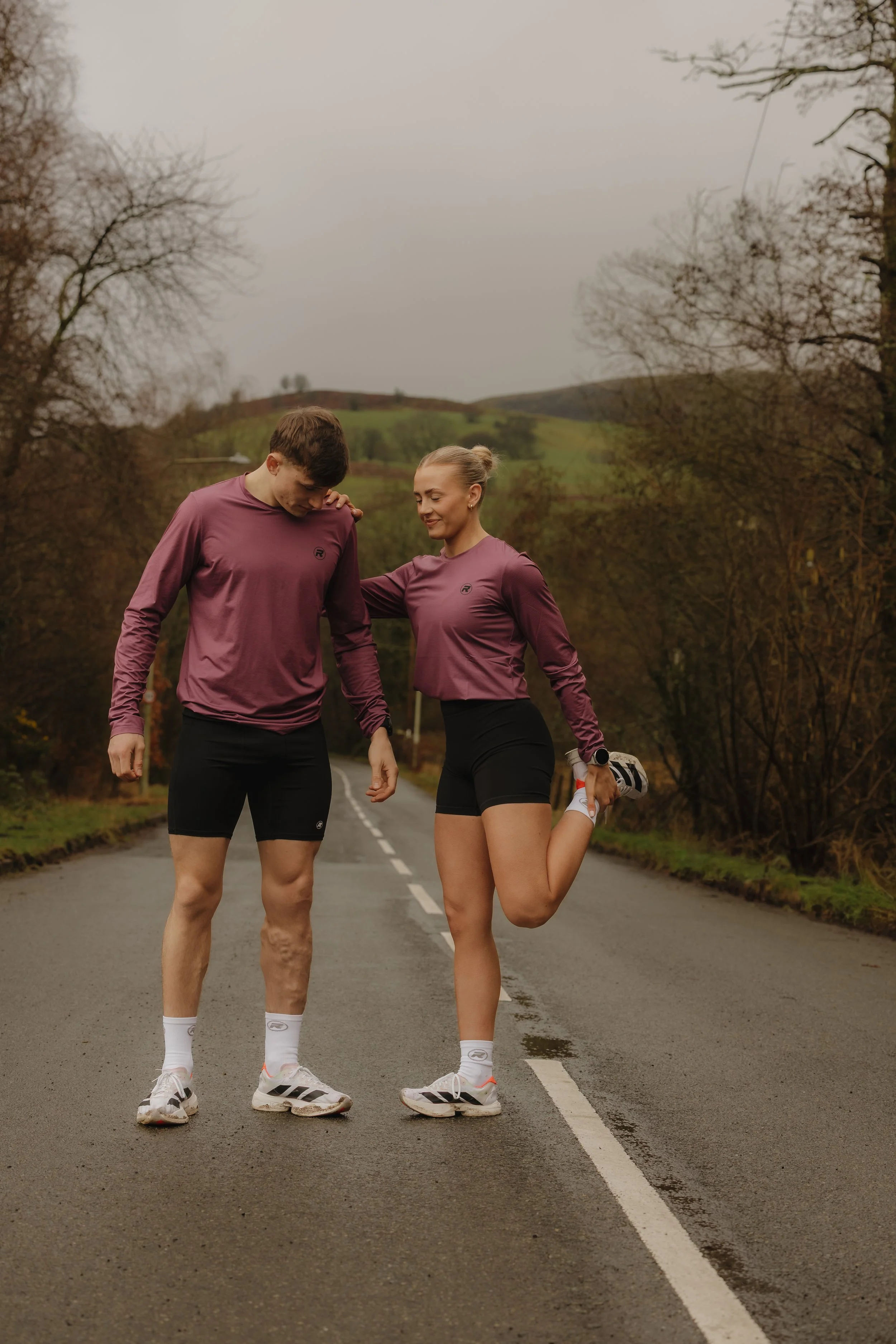 A young man and woman wearing matching purple athletic shirts and black shorts stretching on a rural road with trees and hills in the background.
