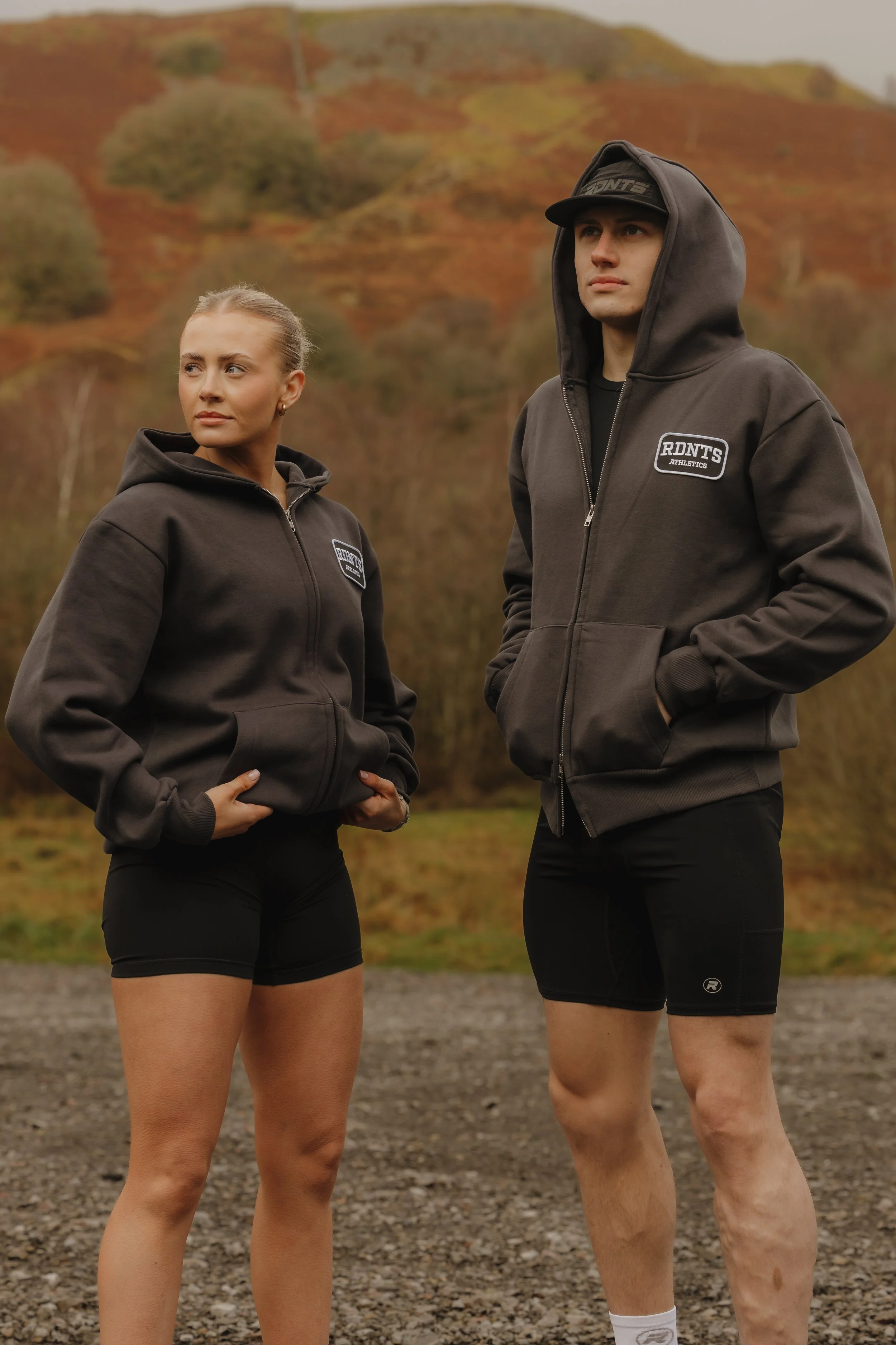 A woman and a man wearing matching black athletic hoodies and shorts standing outdoors on a gravel path, with autumn trees and hills in the background.