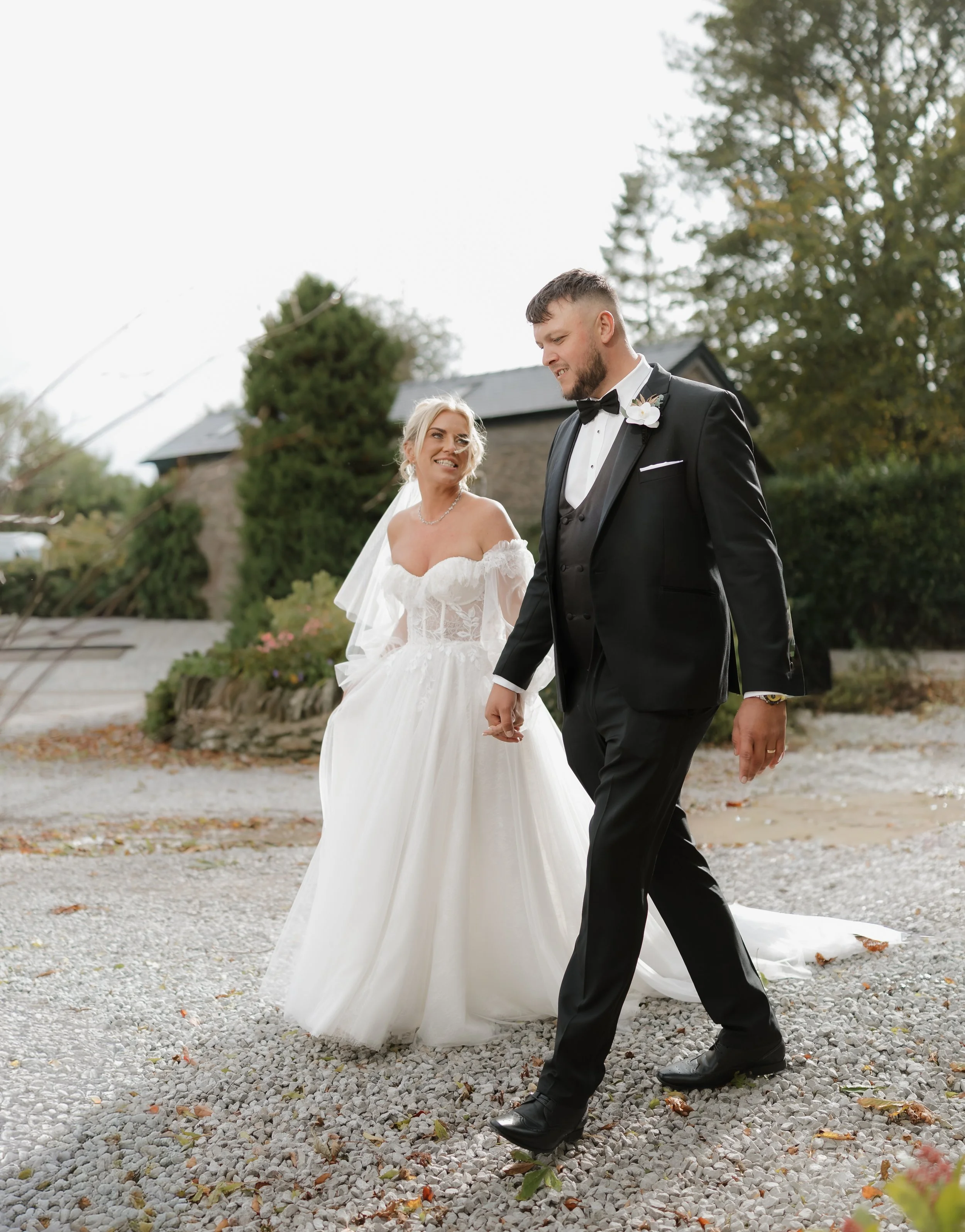 A bride and groom walking outdoors on a gravel path, holding hands, looking at each other, with the bride in a white wedding dress and the groom in a black tuxedo.