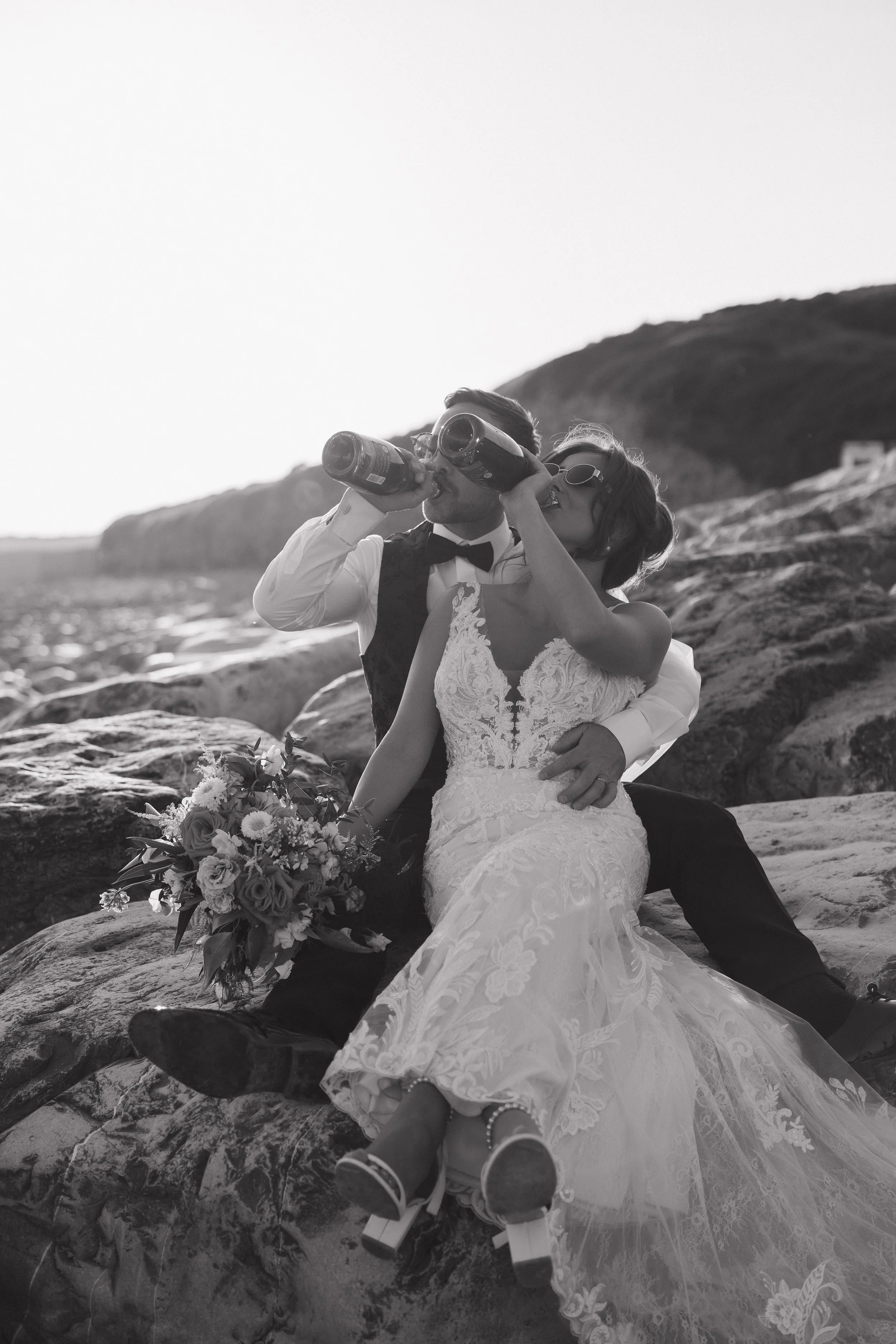 Black and white photo of a newlywed couple sitting on rocks near the beach, drinking from bottles, with the bride holding a bouquet of flowers, and the groom in a tuxedo and bow tie.