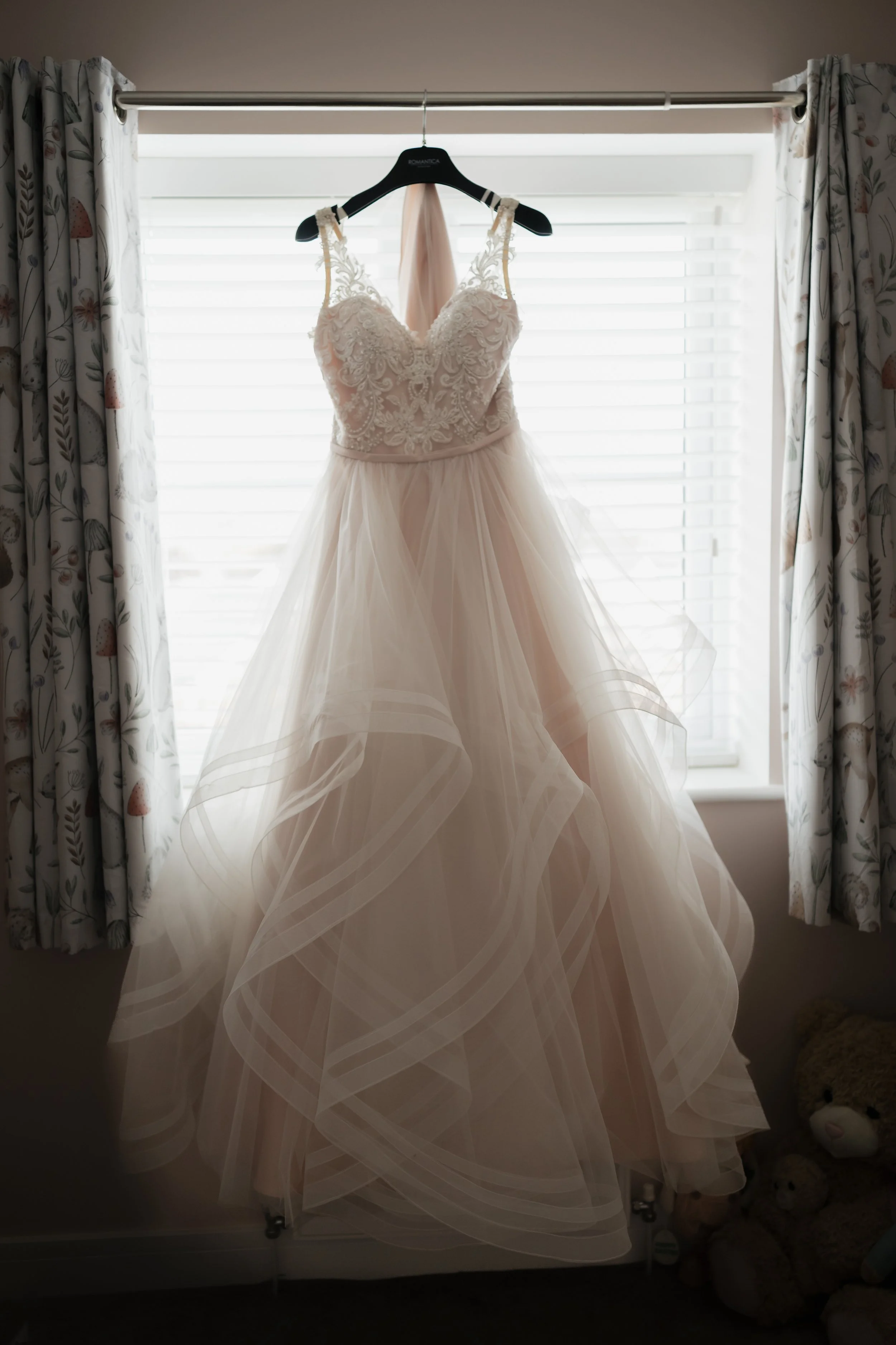 Elegant wedding dress hanging in front of a window with floral curtains.