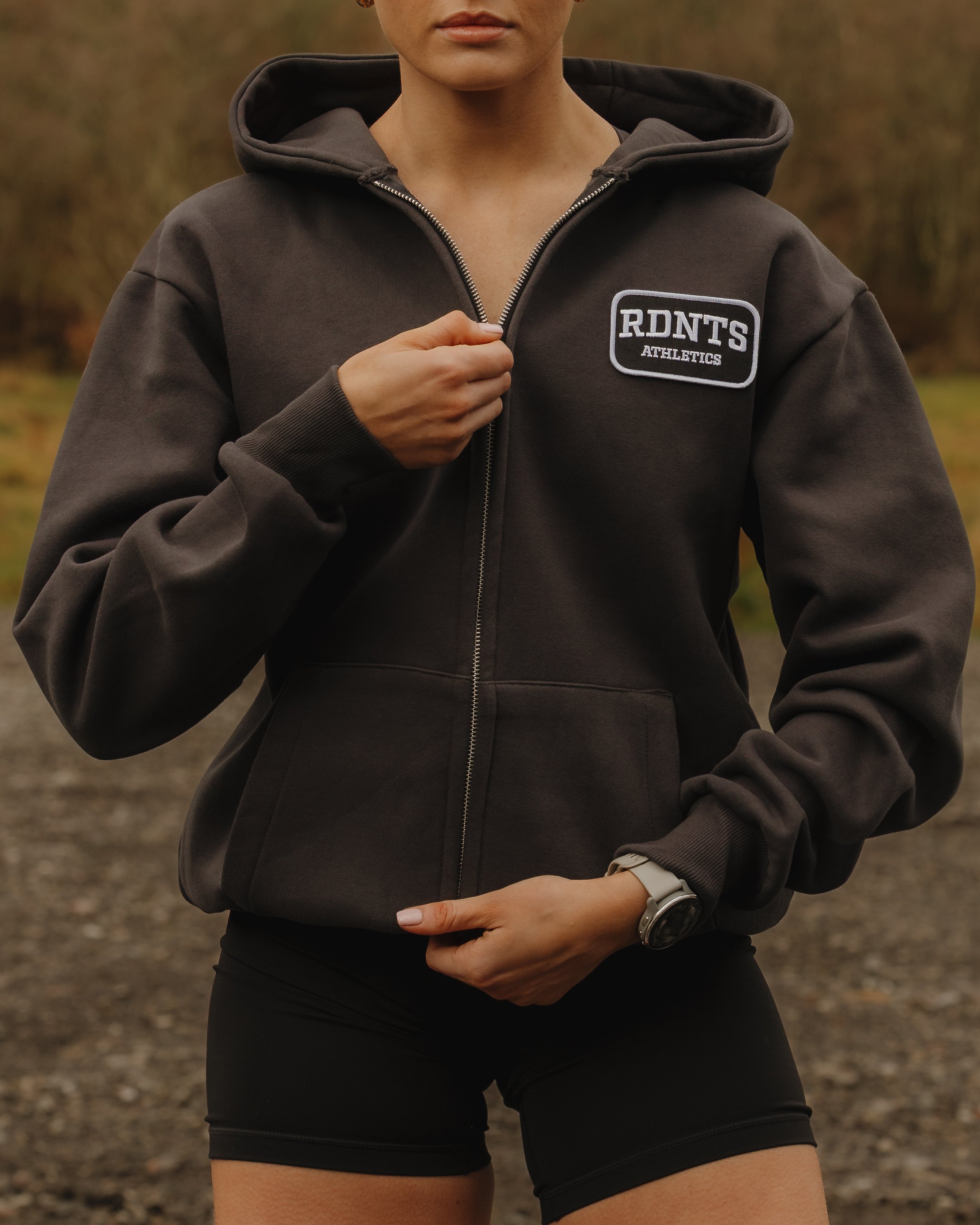 A woman wearing a dark gray hoodie with a patch that reads 'RDNT'S ATHLETICS' and black shorts, outdoors with trees in the background.