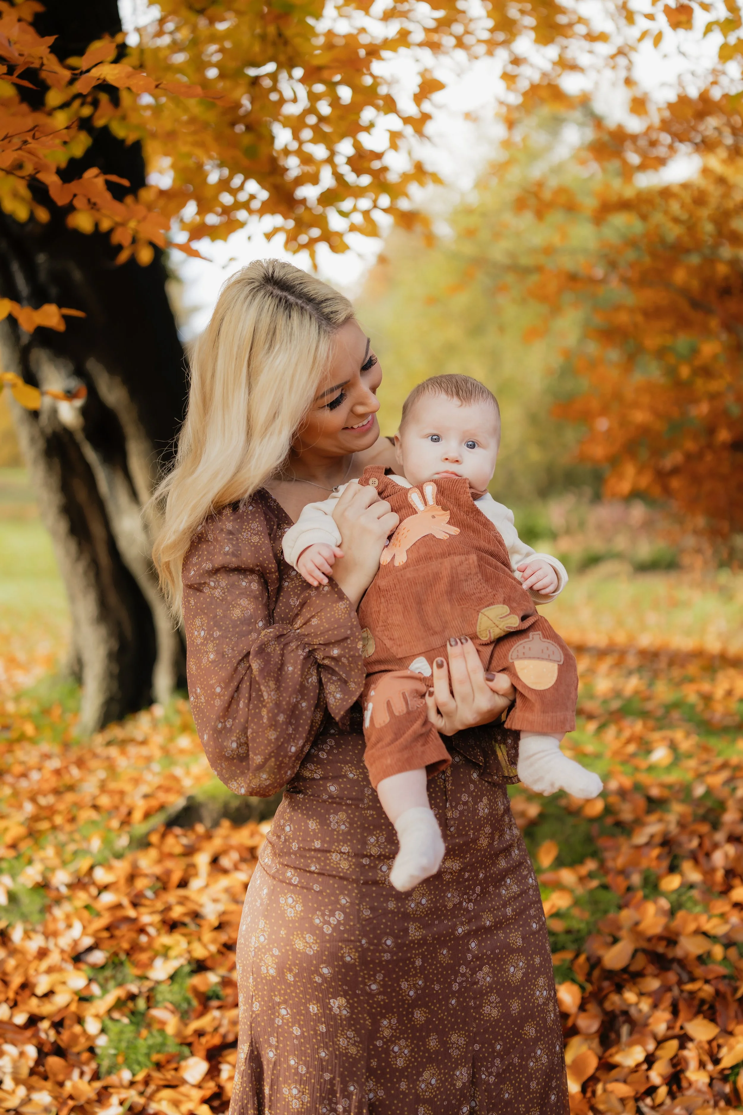 A woman holding a baby outdoors among autumn trees with orange leaves.