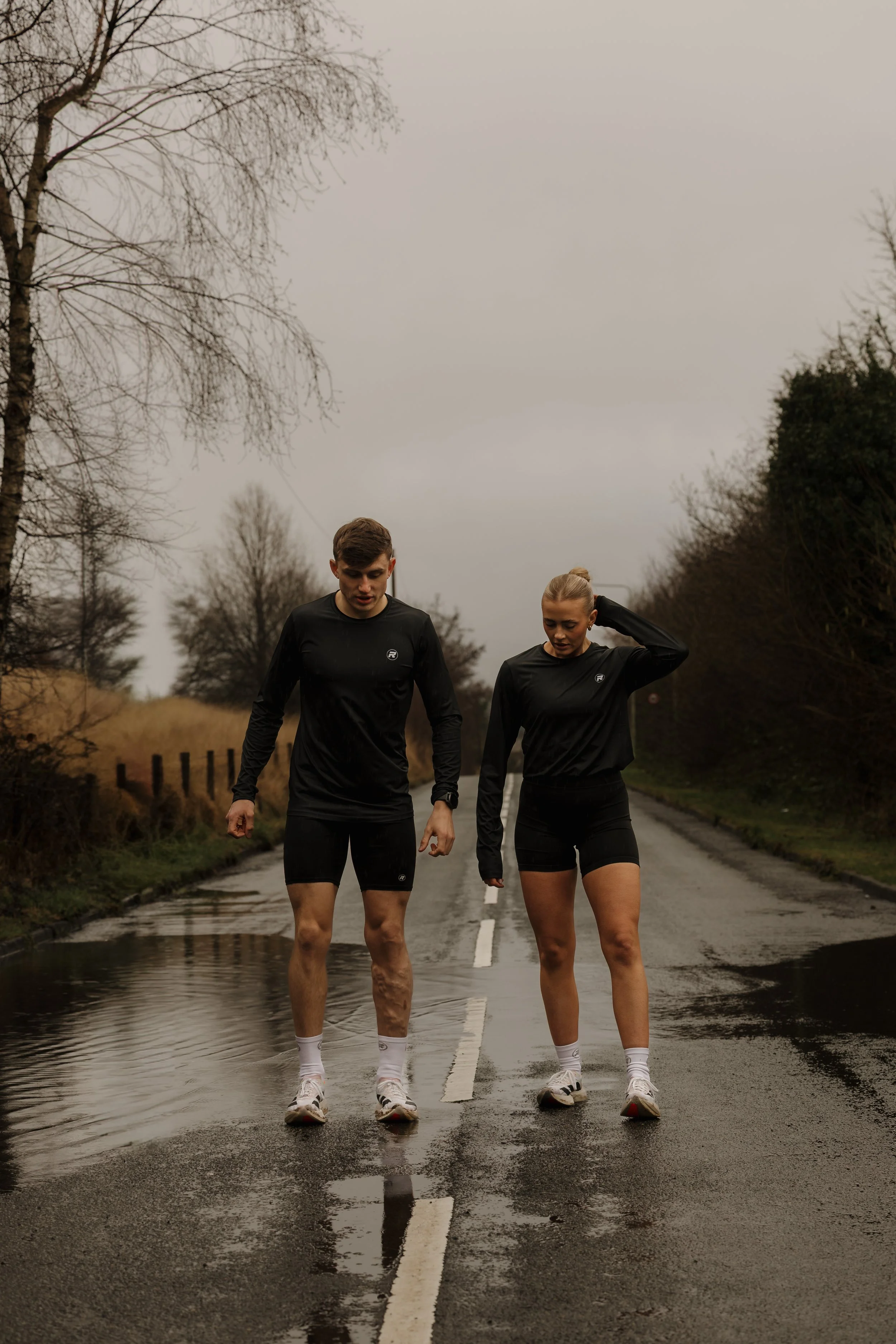 A man and a woman walking together on a flooded road while wearing black athletic clothing, with trees on both sides and an overcast sky.
