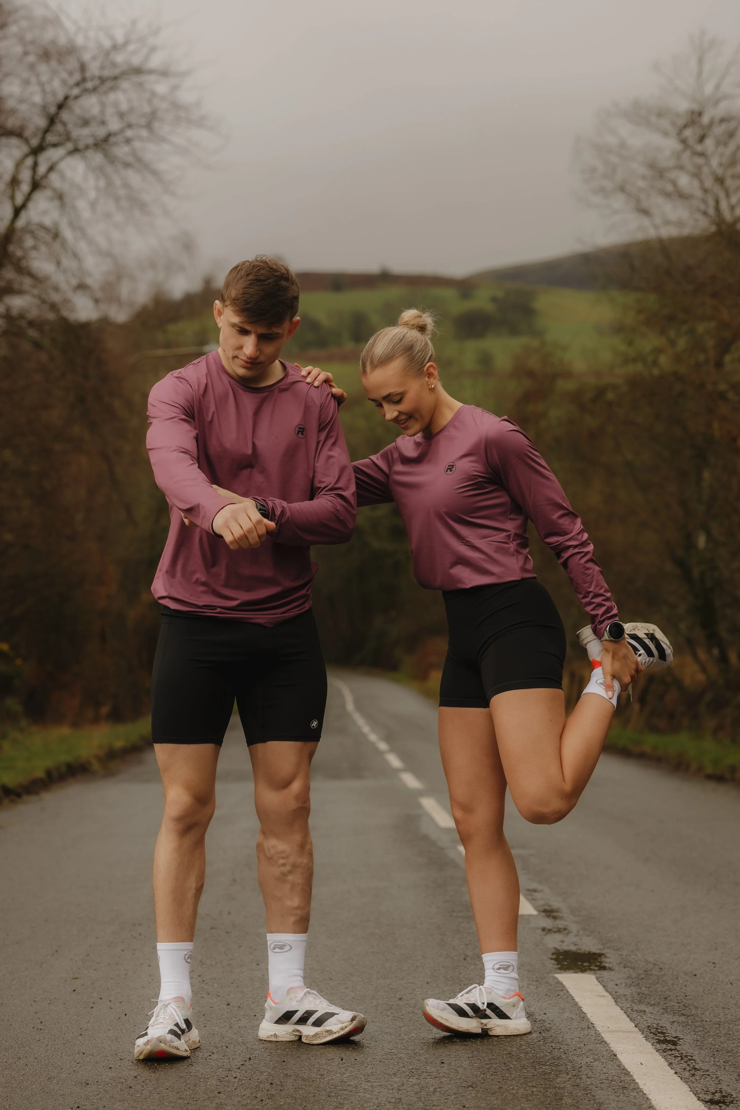A man and a woman in athletic clothing standing on a rural road with trees and hills in the background. The woman is stretching her leg while the man is checking his watch, possibly before or after running.