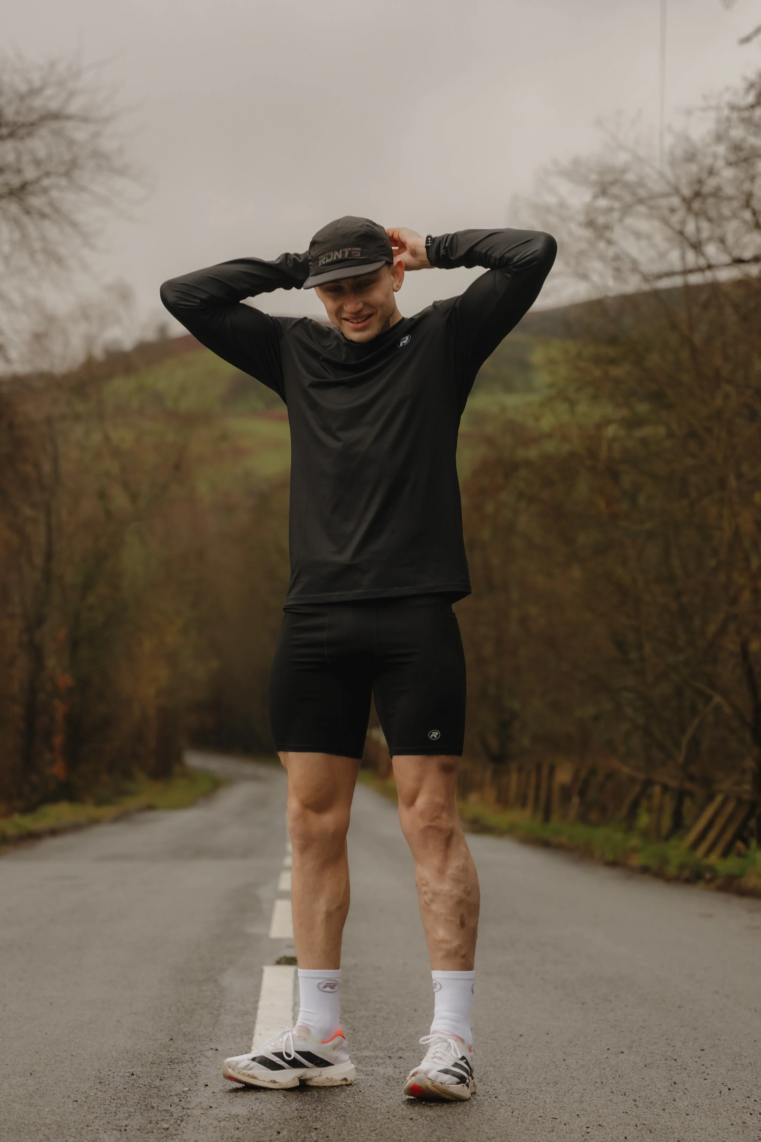 A young man dressed in black athletic gear standing on a rural road, smiling, with a landscape of trees and hills in the background.