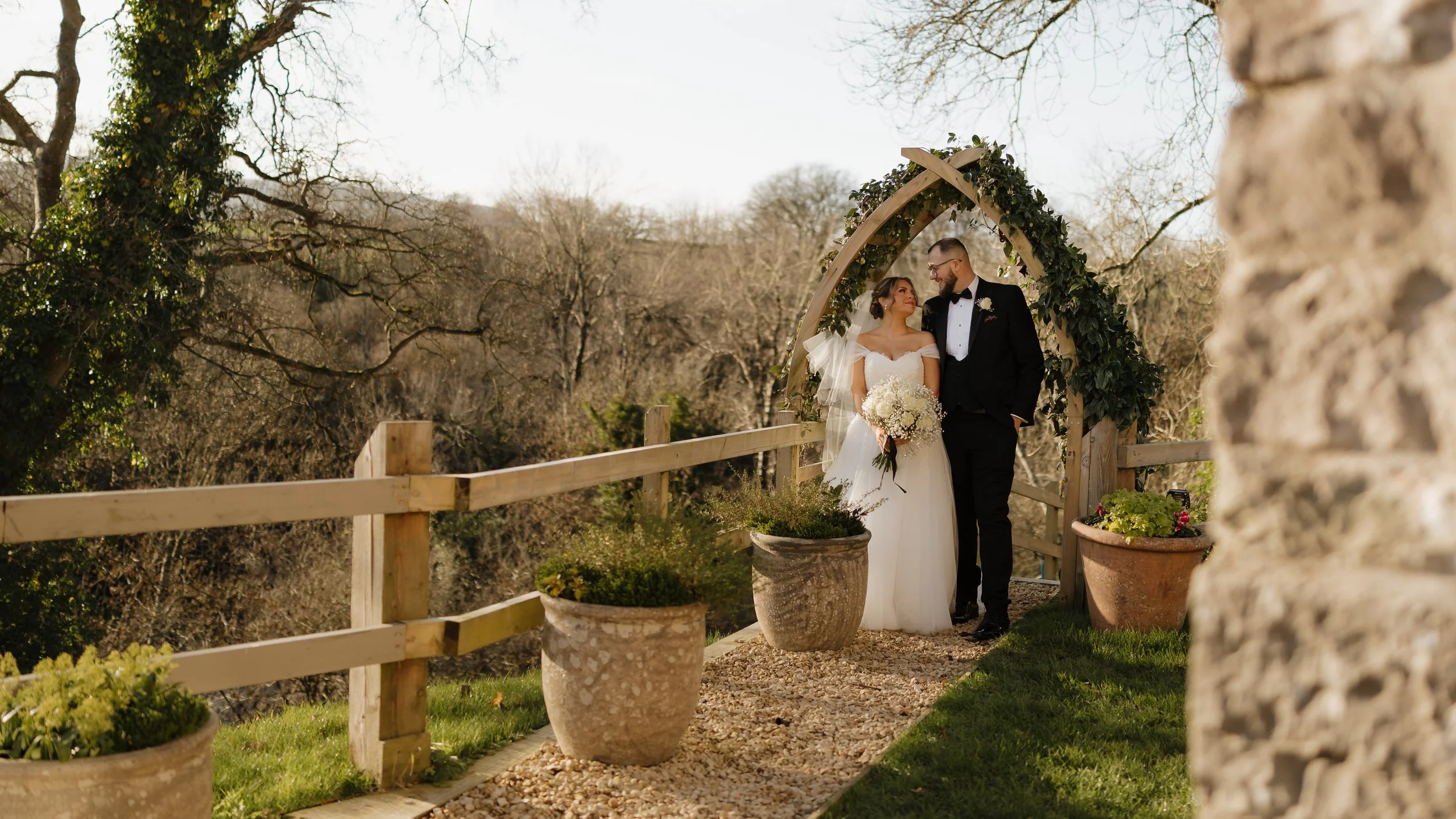 A bride and groom standing under a decorated archway on their wedding day, holding hands and looking at each other, outdoors with trees and a stone wall nearby.