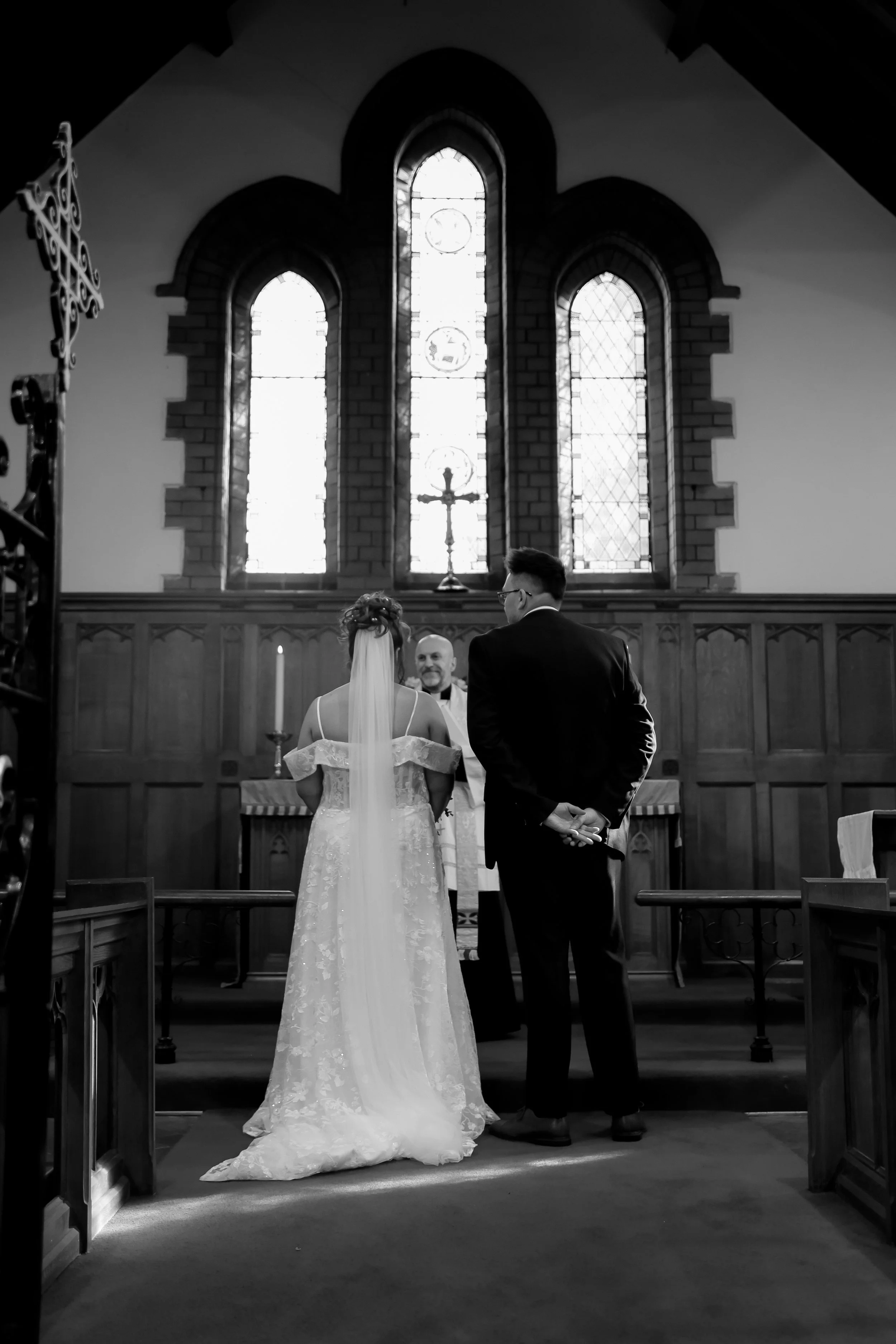 Black and white photo of a wedding ceremony in a church, with a bride, groom, and officiant facing the altar. The bride is in a lace wedding dress with a veil, and the groom is in a dark suit with glasses. The church features tall stained glass windows and wooden paneling.