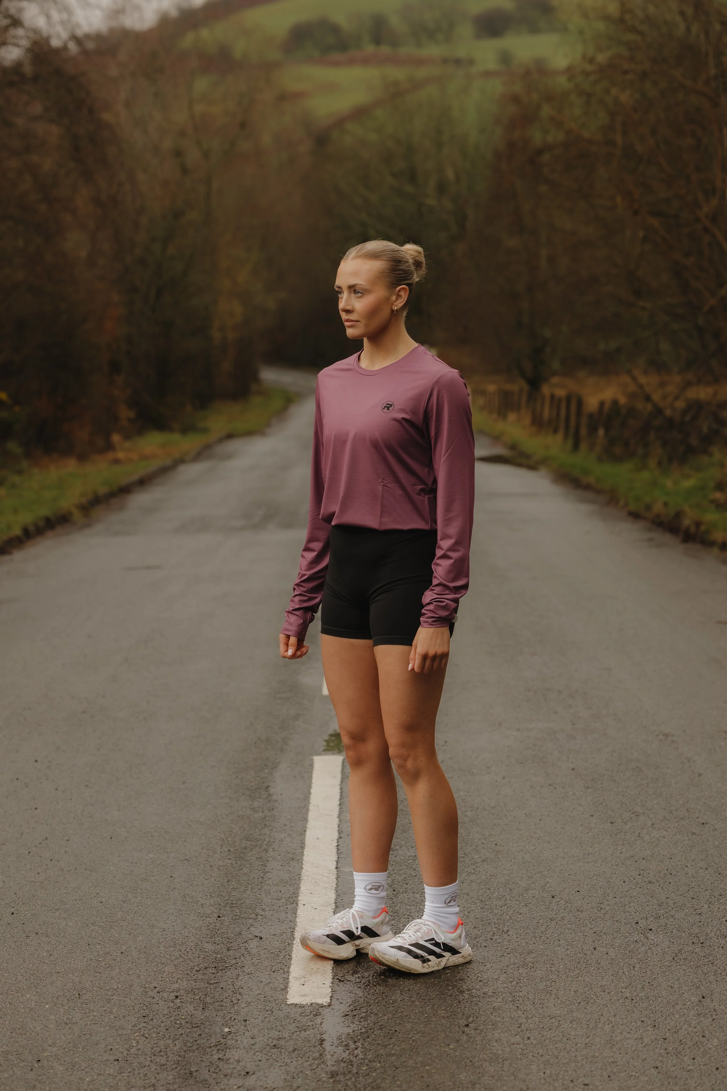 A woman standing alone on a deserted road surrounded by trees and hills, dressed in athletic clothing, including a mauve long-sleeve top, black shorts, white socks, and running shoes.