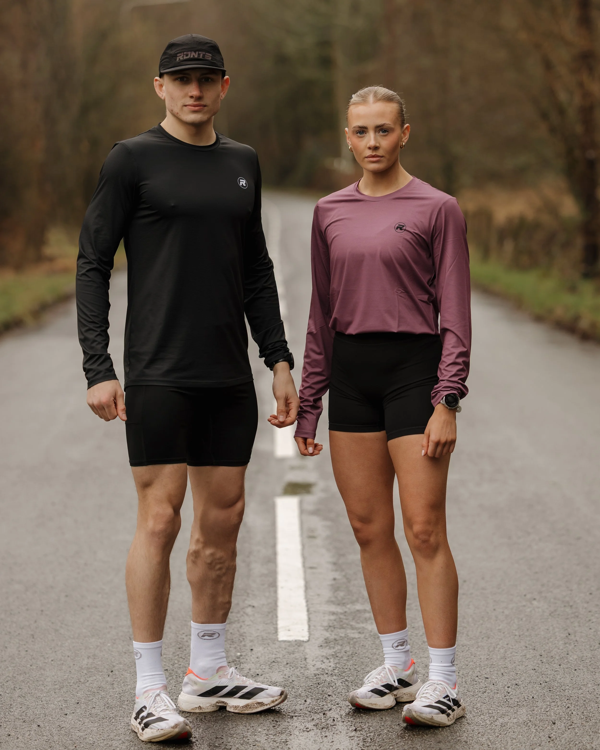 A man and woman standing on an empty rural road, dressed in athletic clothing for running or jogging.