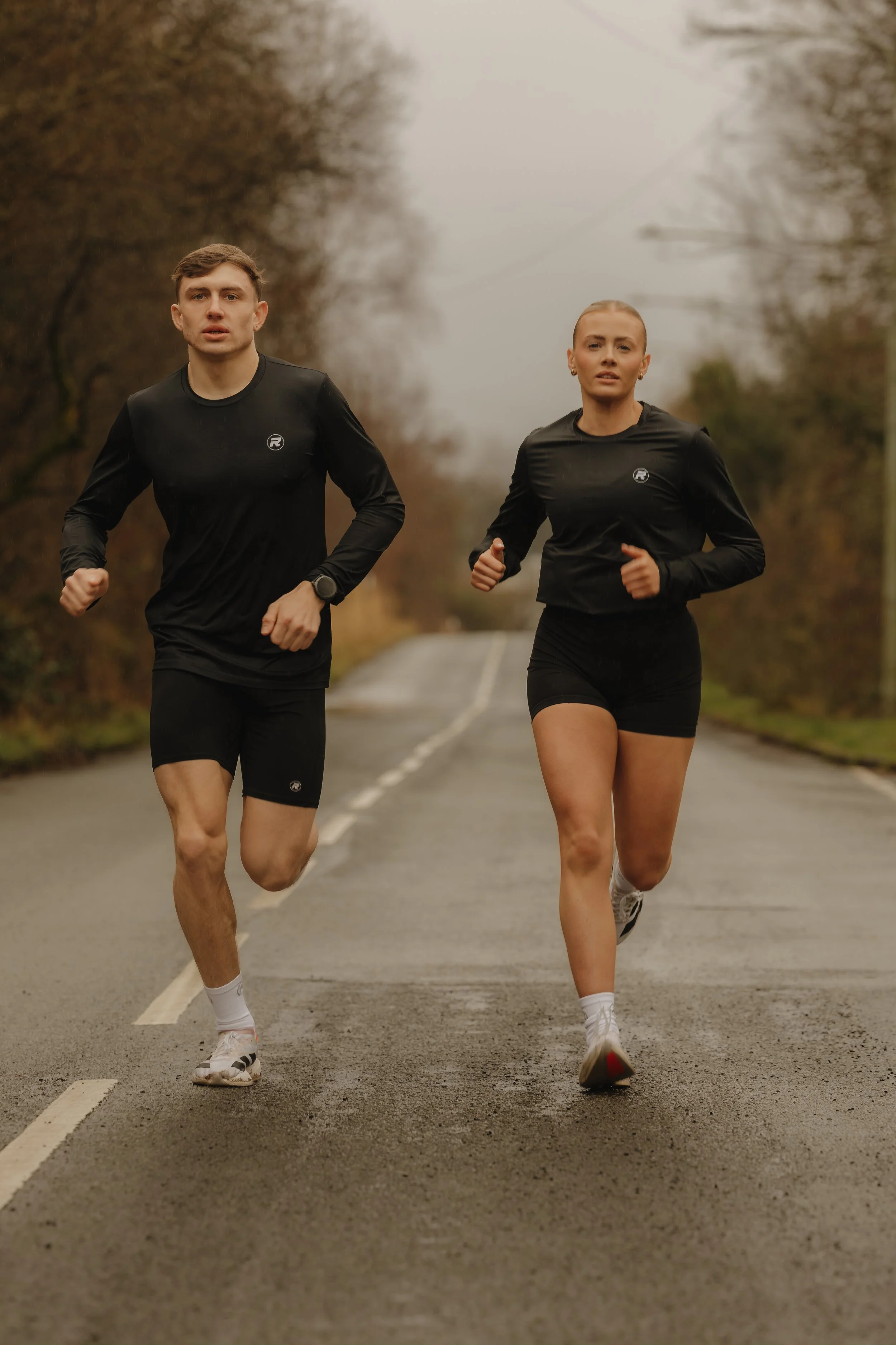 Two people running on a wet road during cloudy weather, surrounded by trees.
