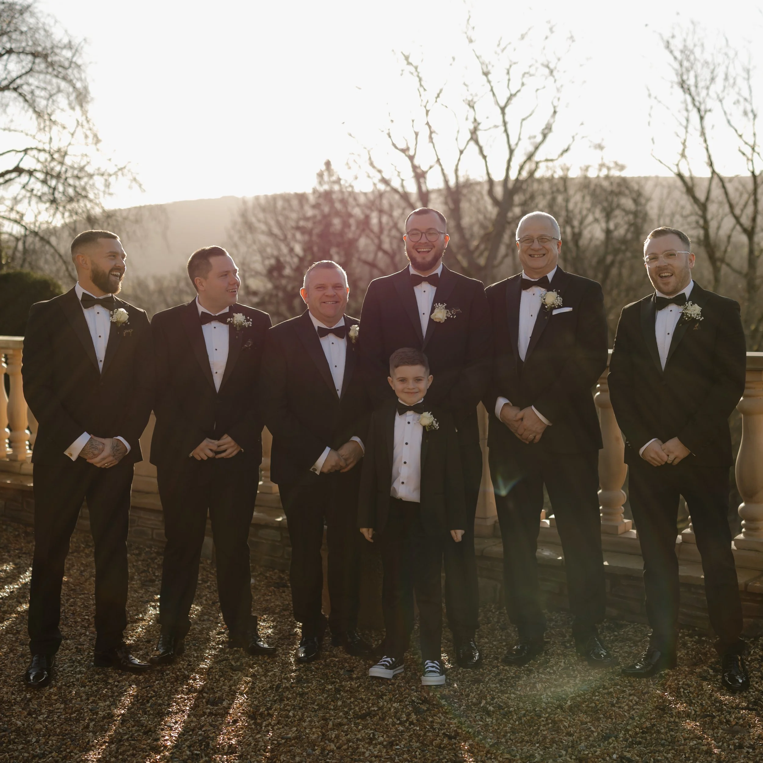 A group of men and a young boy dressed in formal tuxedos standing outdoors, smiling together on a sunny day, with trees and a stone railing in the background.