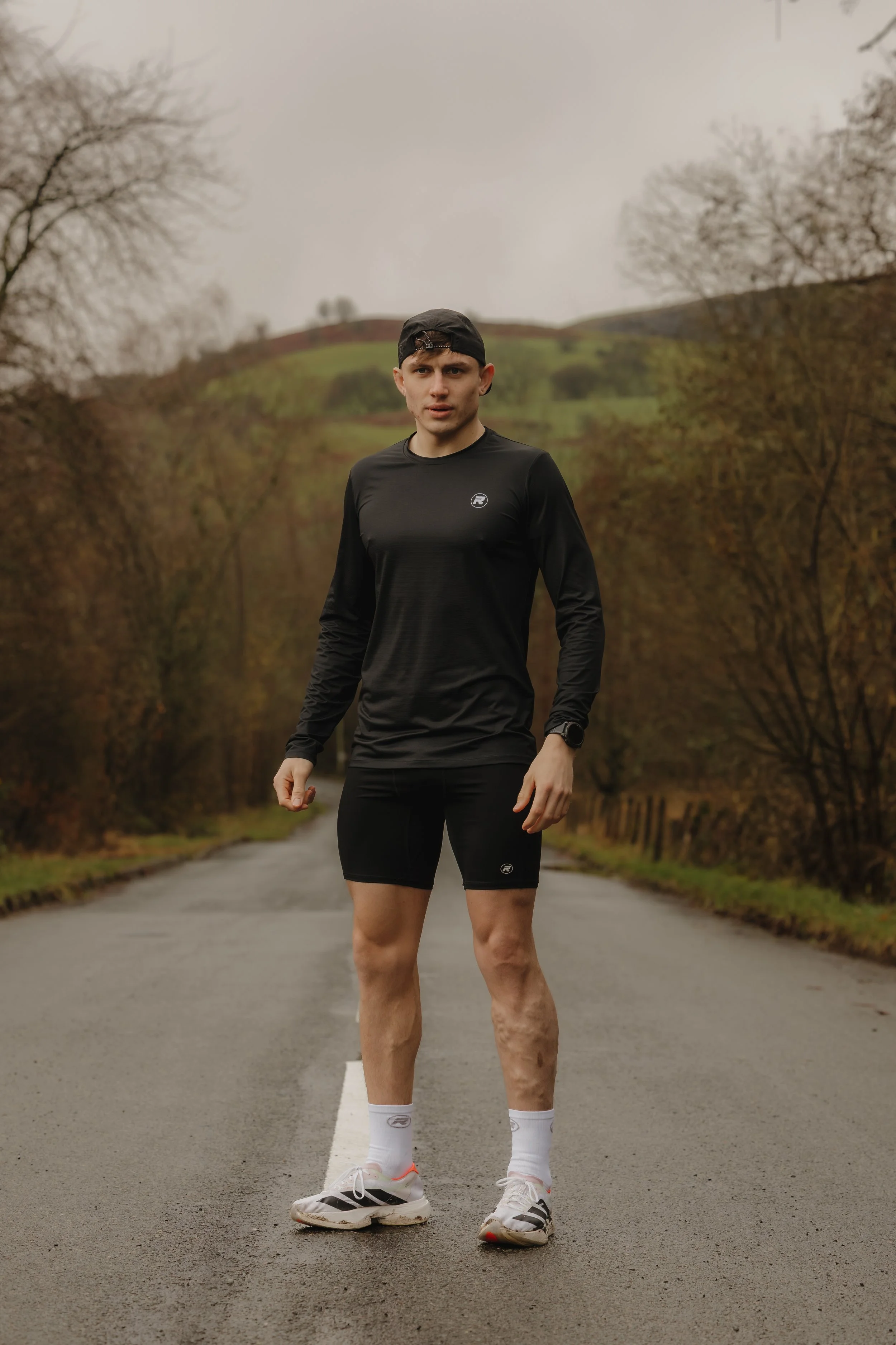 A man running outdoors on a rural road, wearing black athletic clothing and white running shoes, with natural fall scenery in the background.