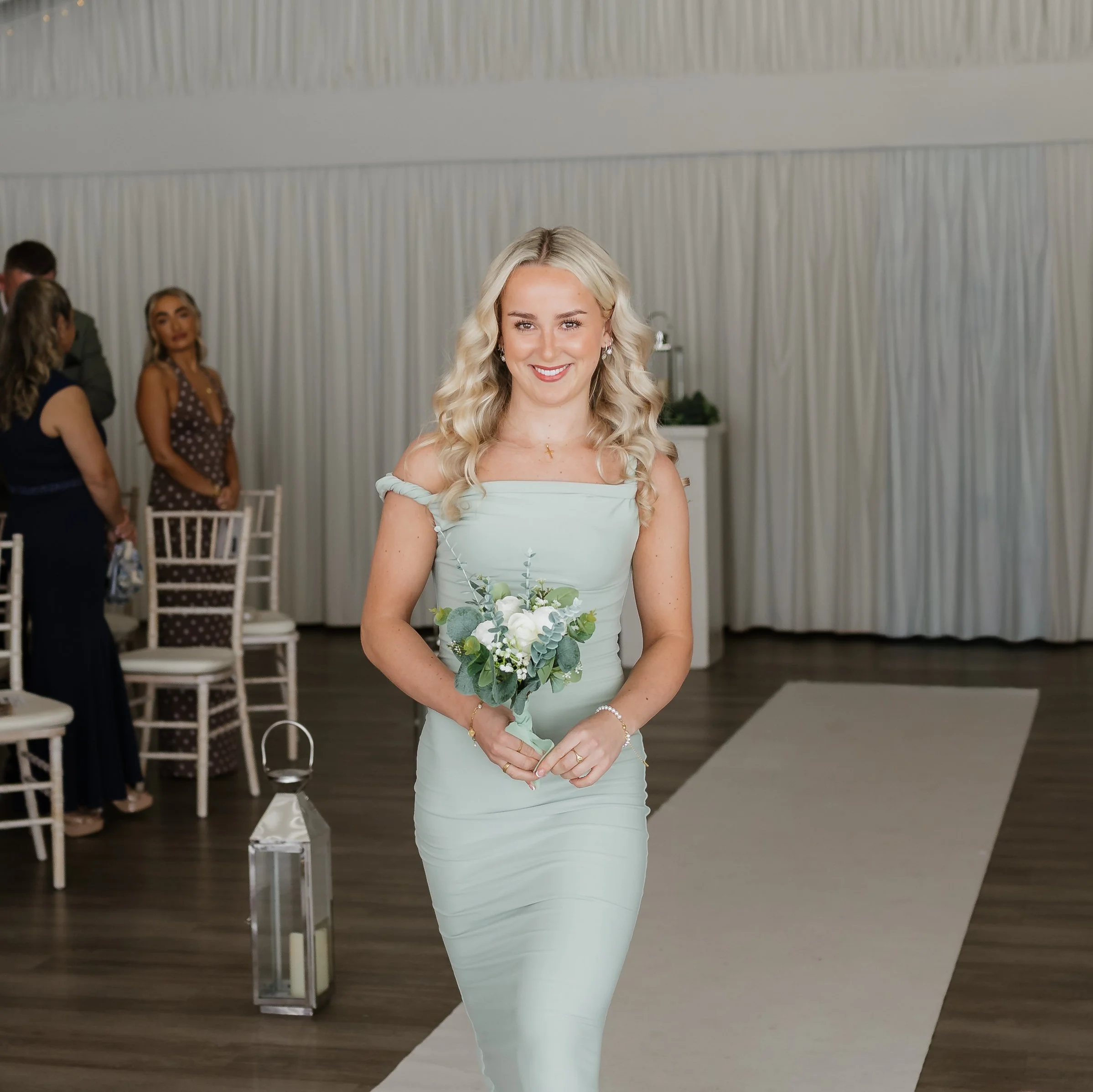 A woman in a pale blue bridesmaid dress holding a bouquet of white flowers, smiling outdoors with autumn-colored trees in the background.