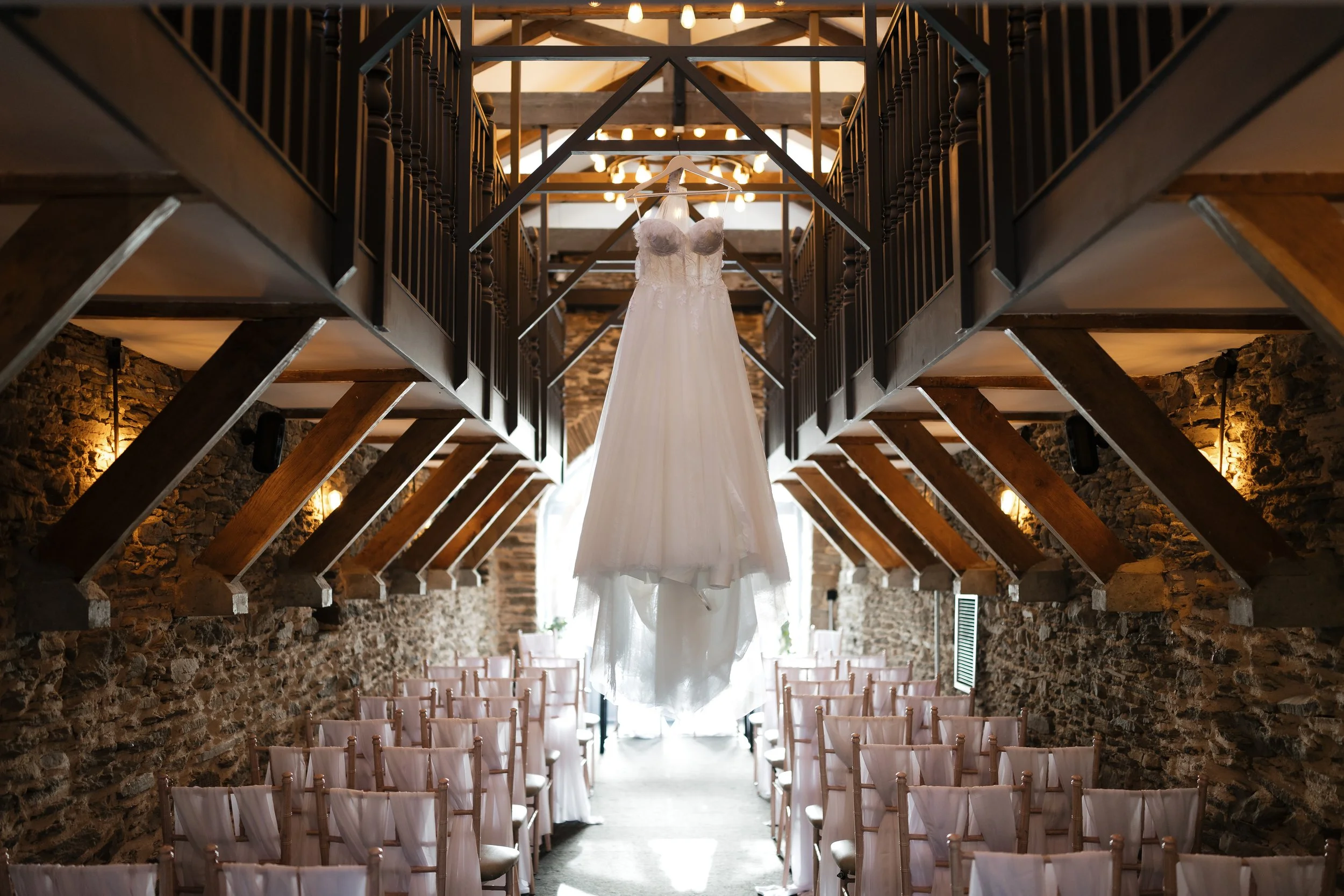 Wedding dress hanging from the ceiling in a rustic wedding venue with wooden beams, stone walls, and rows of chairs.