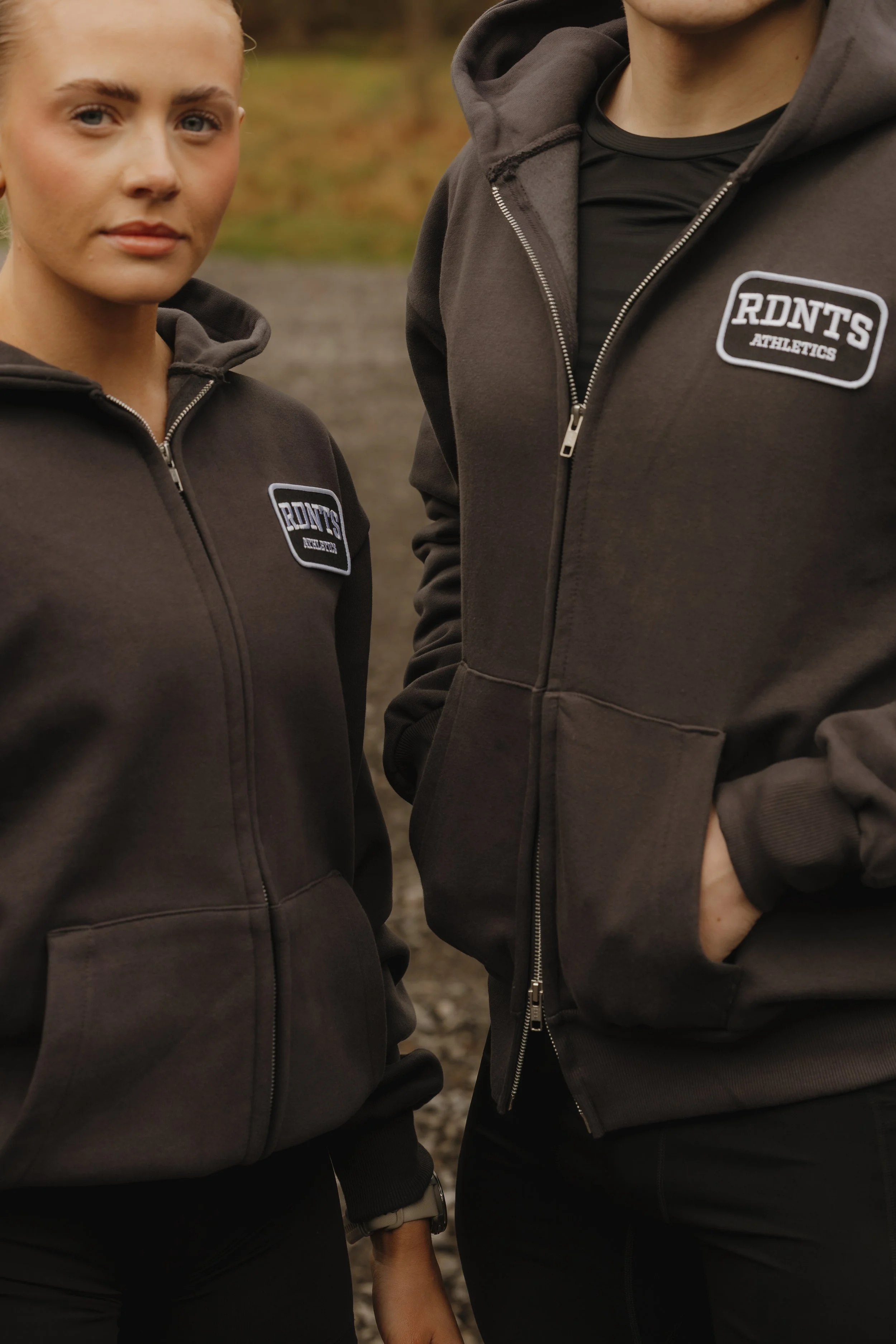 Close-up of two people wearing black hoodies with a logo that says 'RDNT'S ATHLETICS,' standing outdoors.