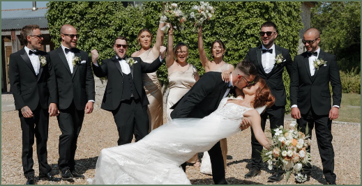 A wedding party outdoors with a bride and groom in front, surrounded by groomsmen and bridesmaids. The bride is wearing a white gown and holding a bouquet, while the groom is in a black tuxedo, as are the groomsmen. The wedding party is celebrating, with some members raising their hands and smiling.