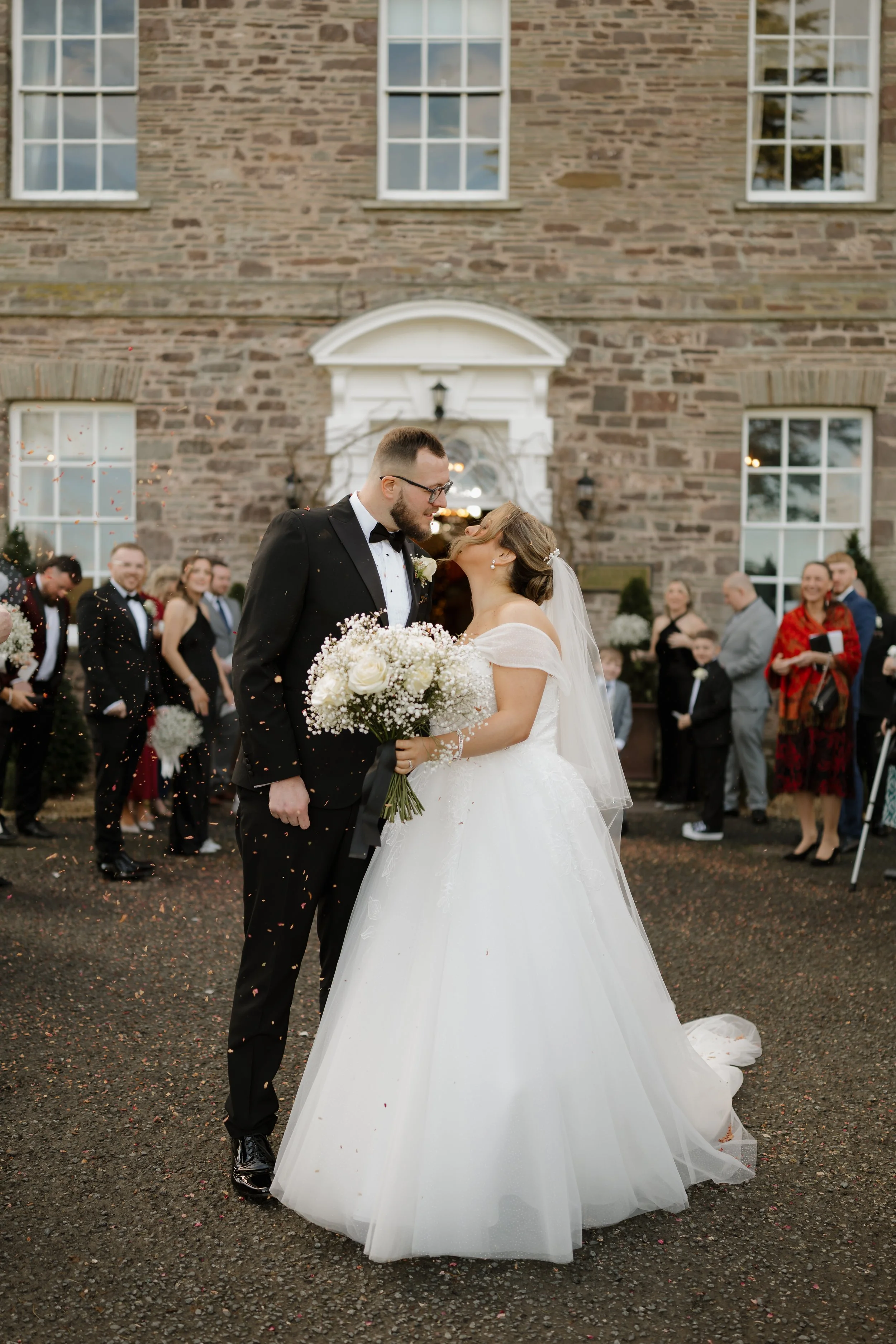 Bride and groom sharing a kiss at their wedding ceremony outside a brick building, with guests watching and throwing confetti.