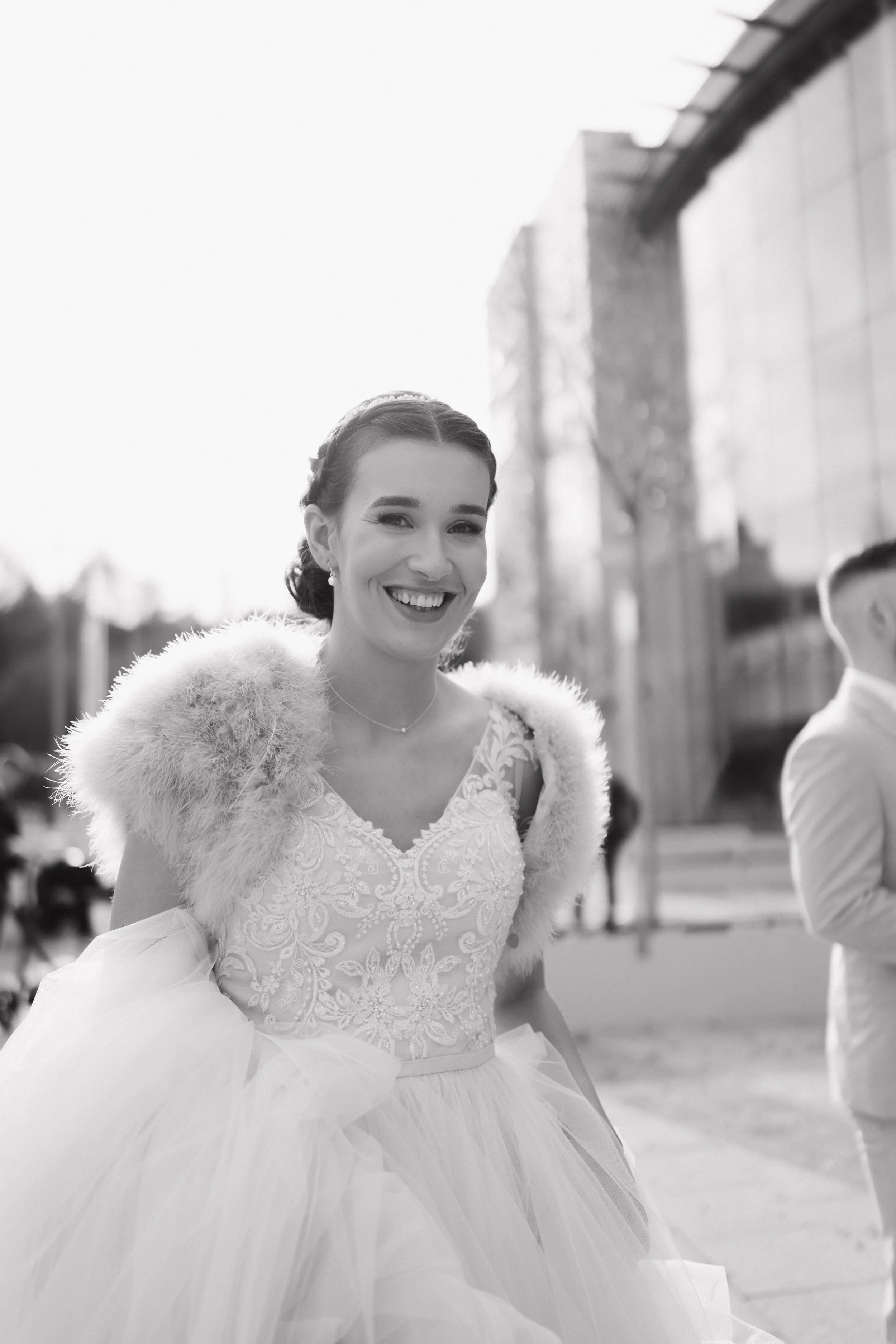 A woman in a white wedding dress with lace embroidery and a fur shawl, smiling outdoors in black and white.