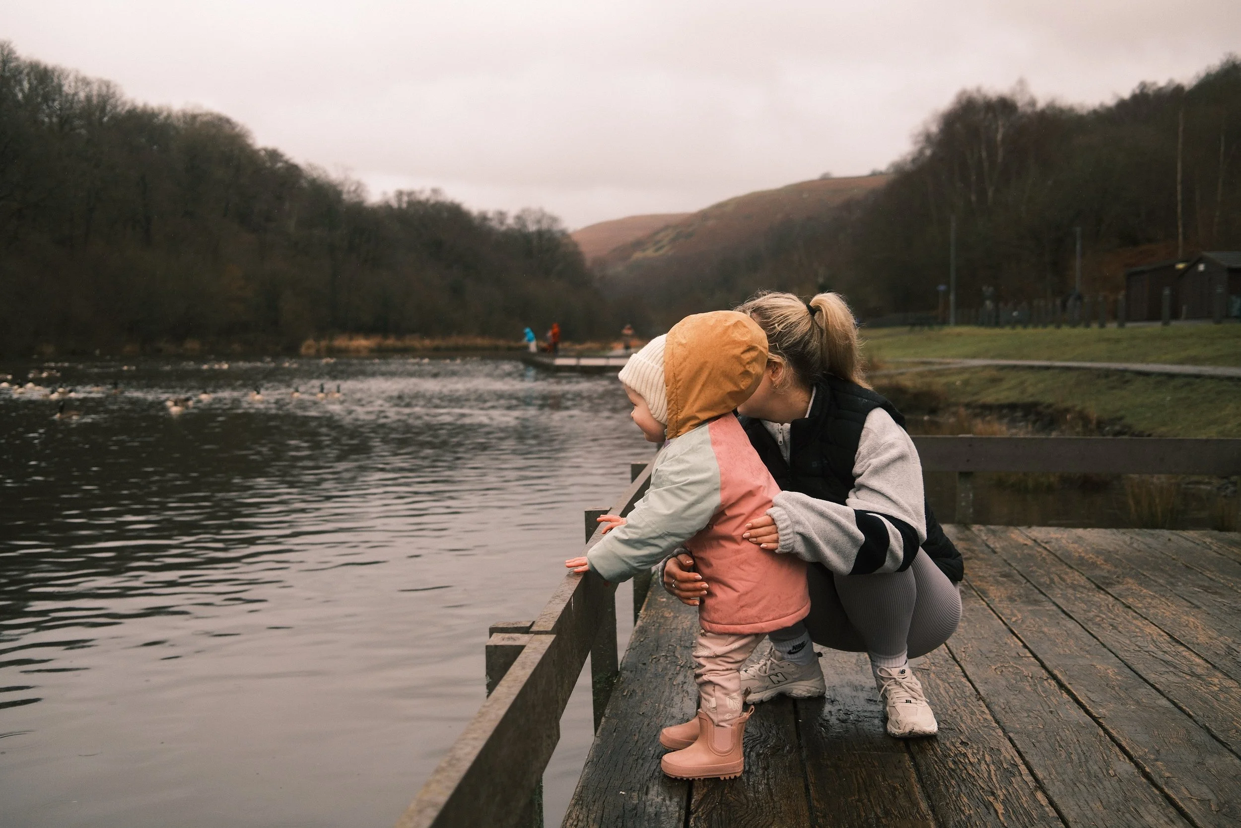A woman and young girl standing on a wooden dock by a river, looking out at the water on a cloudy, overcast day.