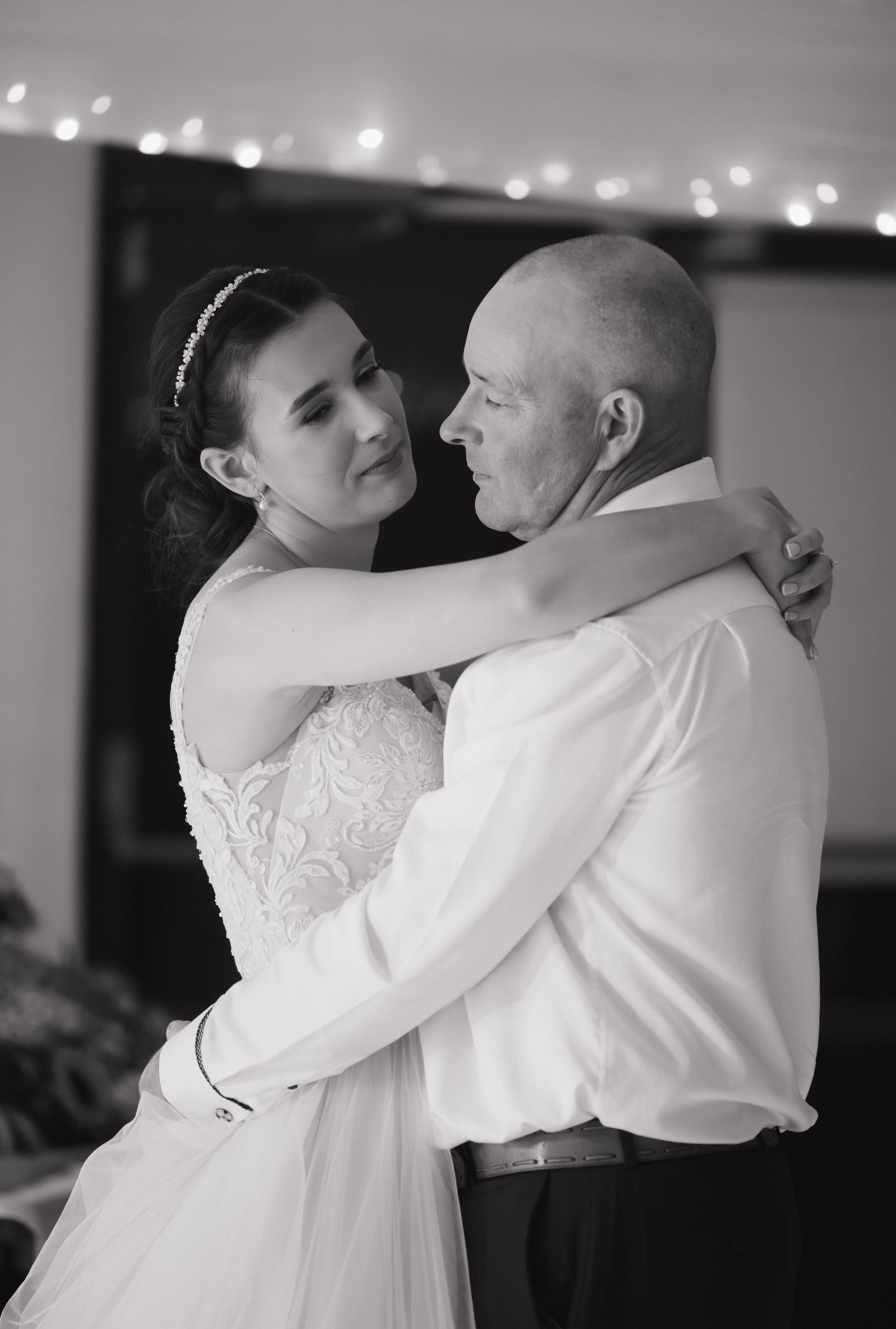 A bride and a man, possibly her father, dance closely at a wedding reception under string lights. The bride wears a lace wedding dress and a headband, and the man is in a white shirt.