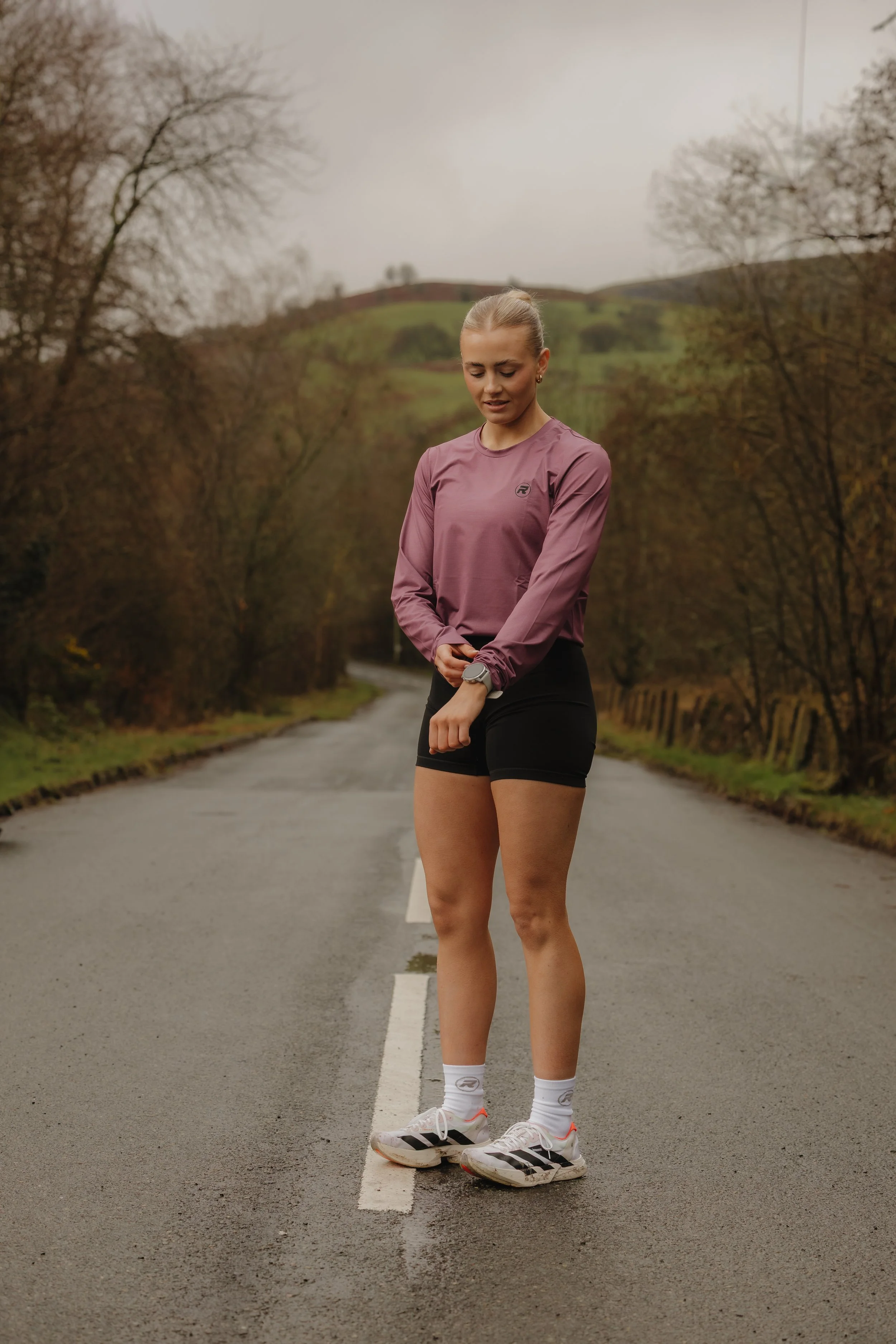 A woman in athletic clothing looking at her watch on a rural road surrounded by trees and hills.