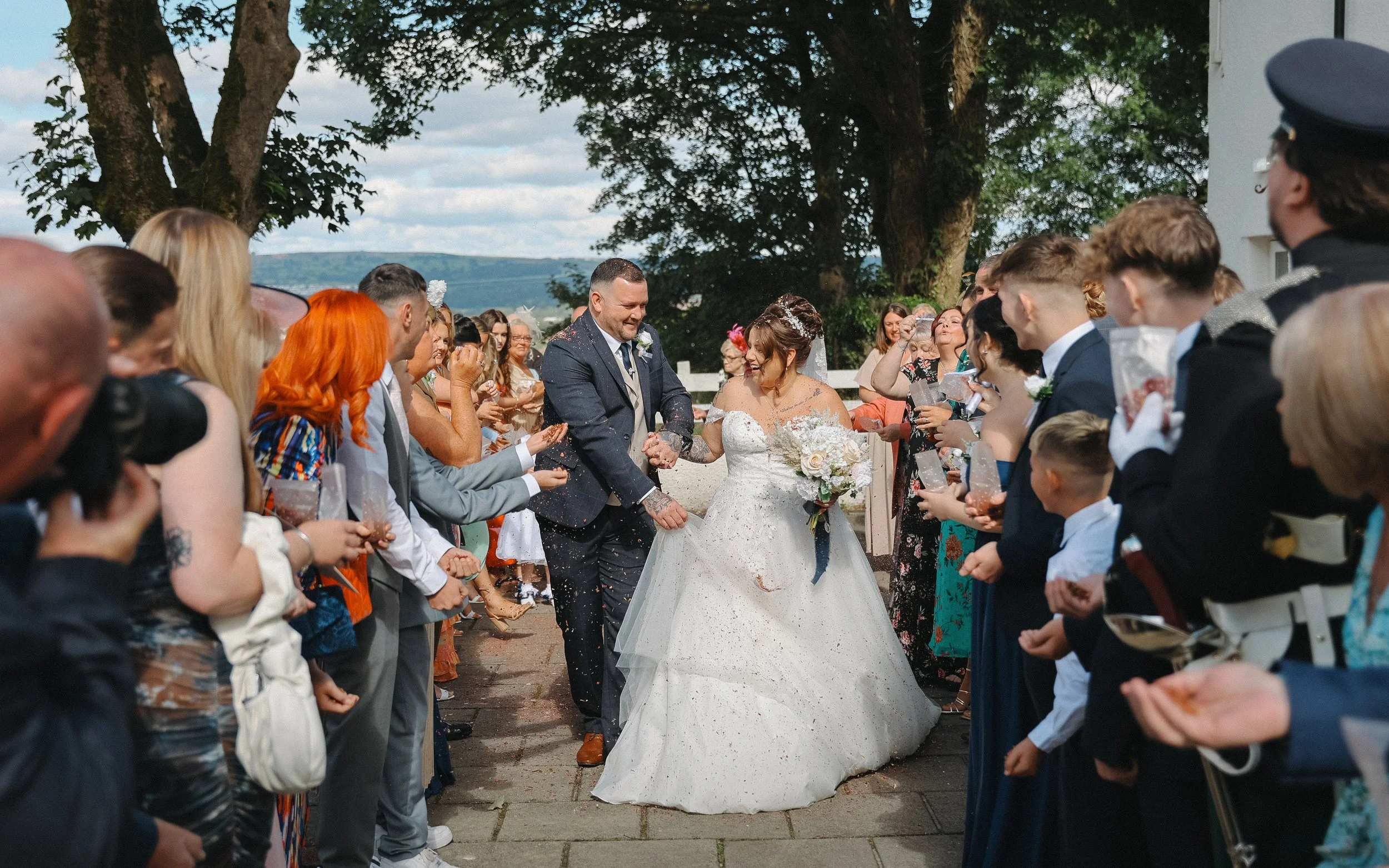 Bride and groom smiling as guests celebrate at a wedding outdoors on a sunny day, with trees in the background.