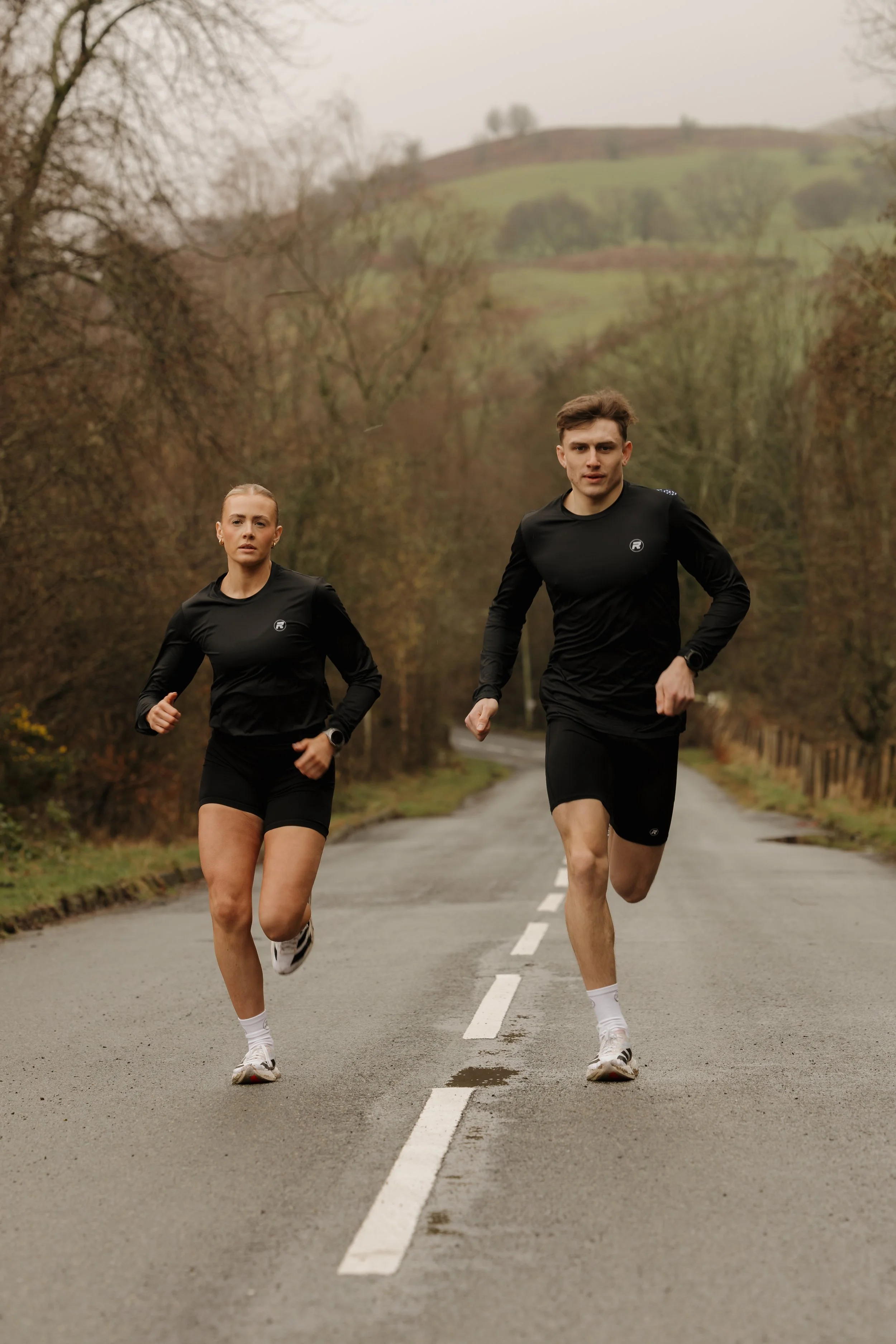 Two runners, a woman and a man, running on a rural road surrounded by trees and hills in the background.