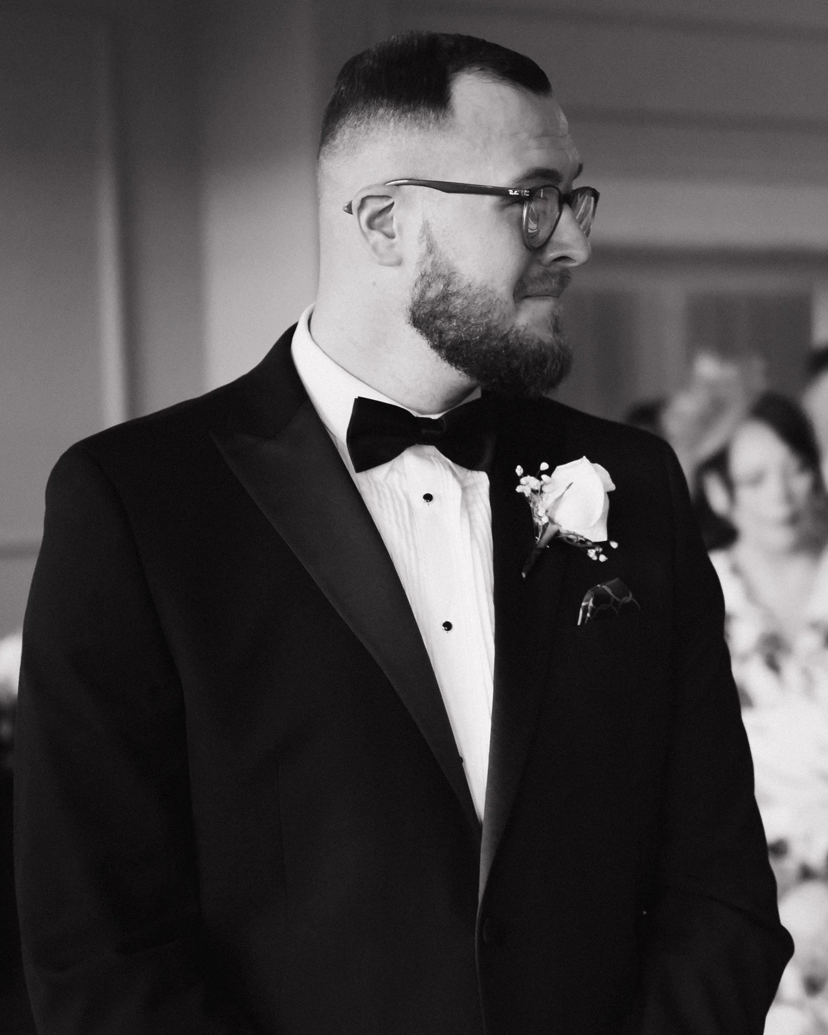 A man in a tuxedo with a bow tie, glasses, and a boutonniere, looking to the side at a formal event.