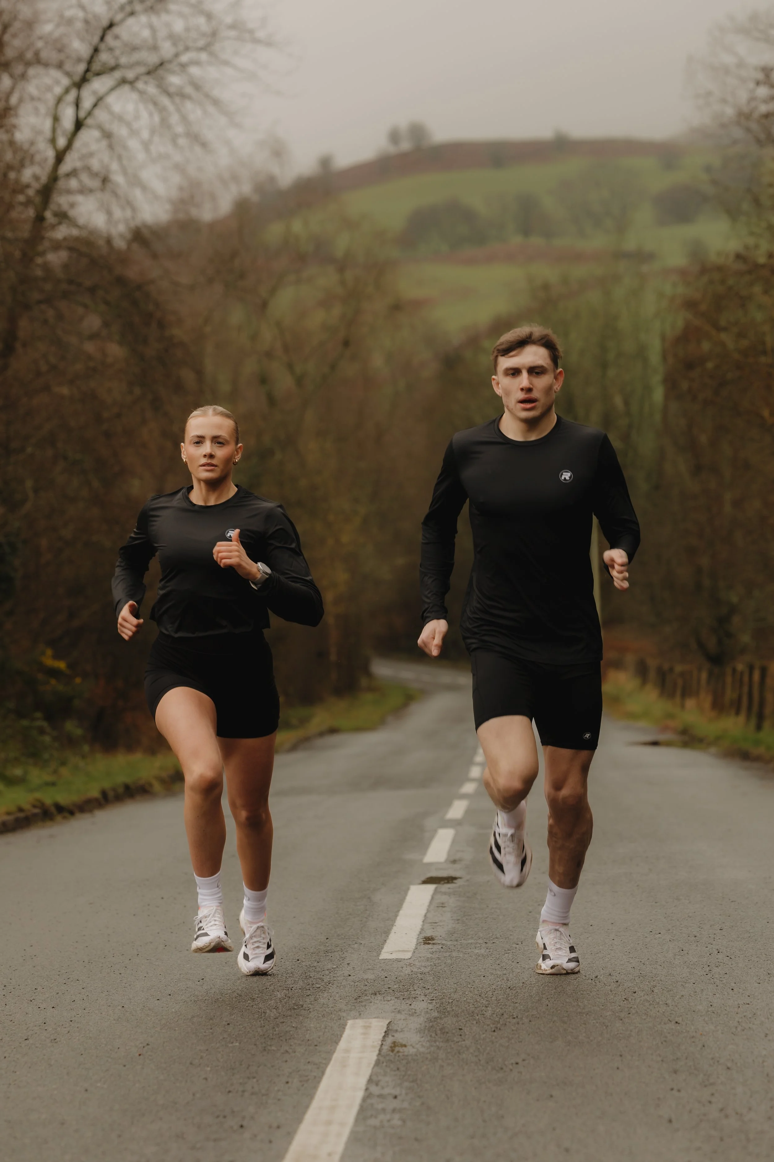 Man and woman running on an empty road surrounded by trees and hills in the background.