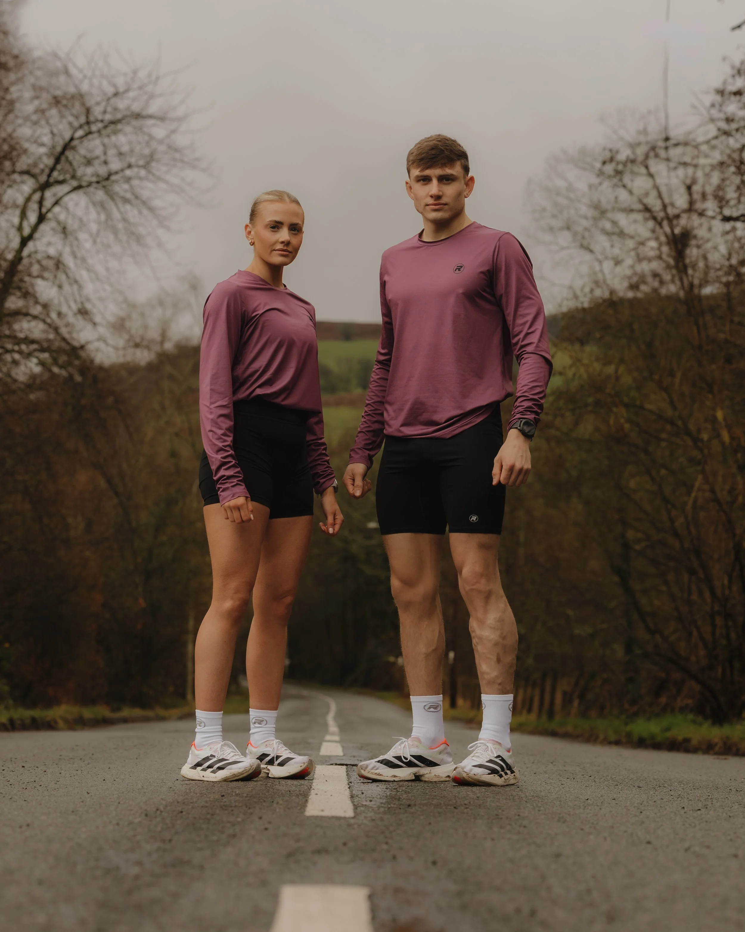 A young man and woman standing on a rural road in athletic wear, facing the camera with a backdrop of trees and overcast sky.