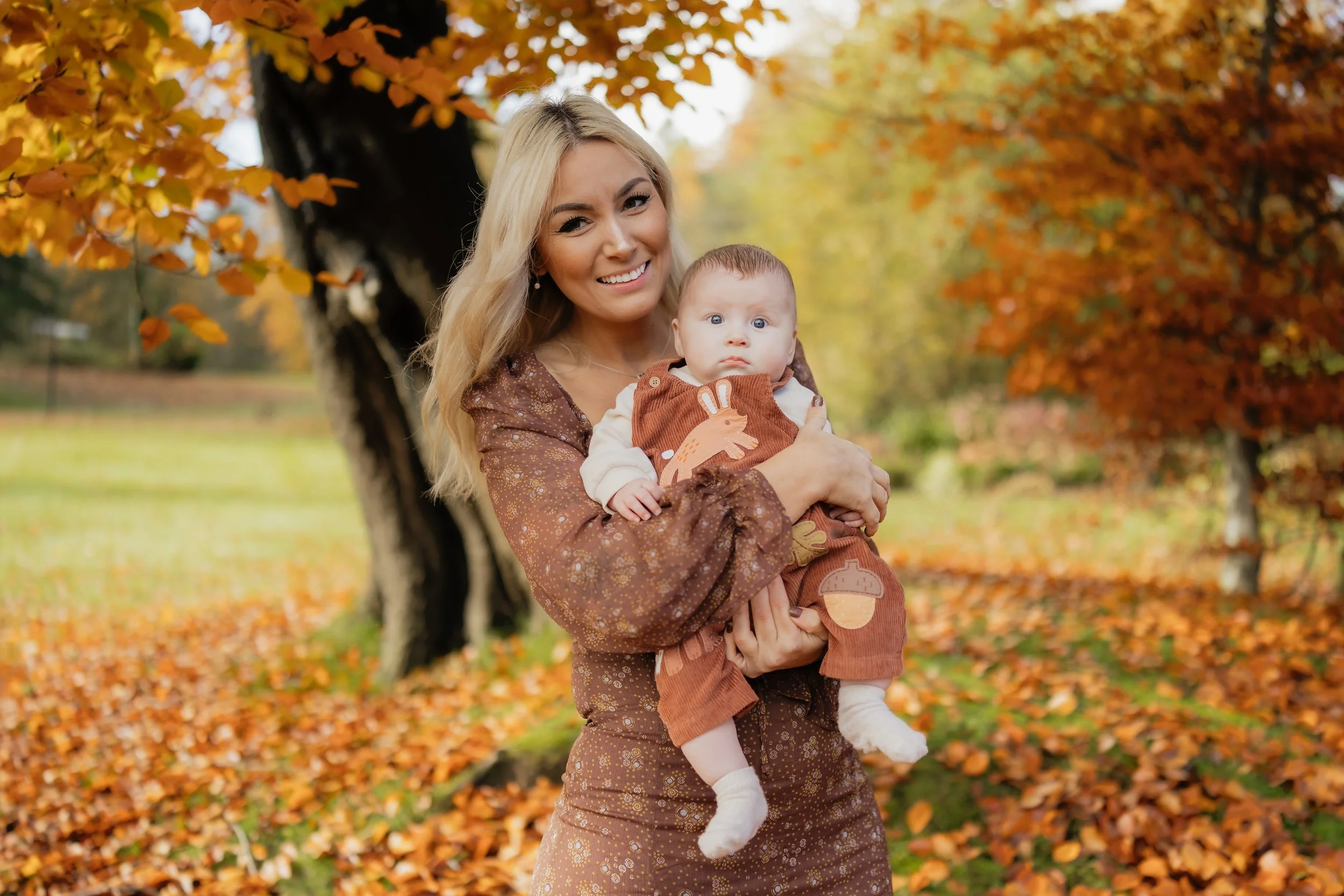 A woman with blonde hair smiles while holding a baby in an autumn park. The woman wears a brown dress with a floral pattern, and the baby wears a brown overalls with a bunny and mushroom design. colourful fall leaves cover the ground and trees with o