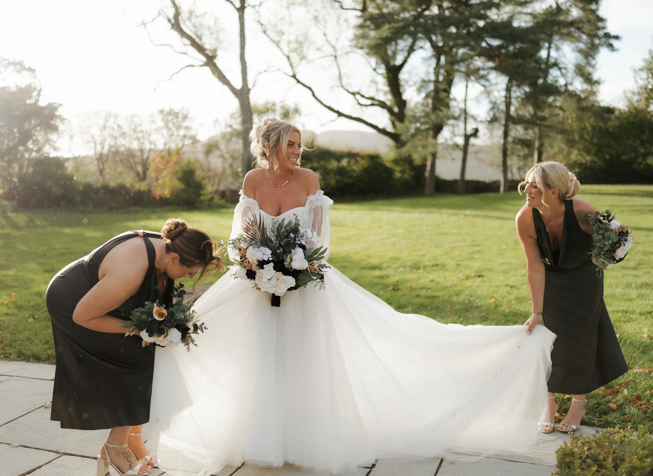A bride in a white wedding dress holding a bouquet is smiling and playing with two bridesmaids in black dresses outdoors on a sunny day, on a grassy lawn with trees in the background.