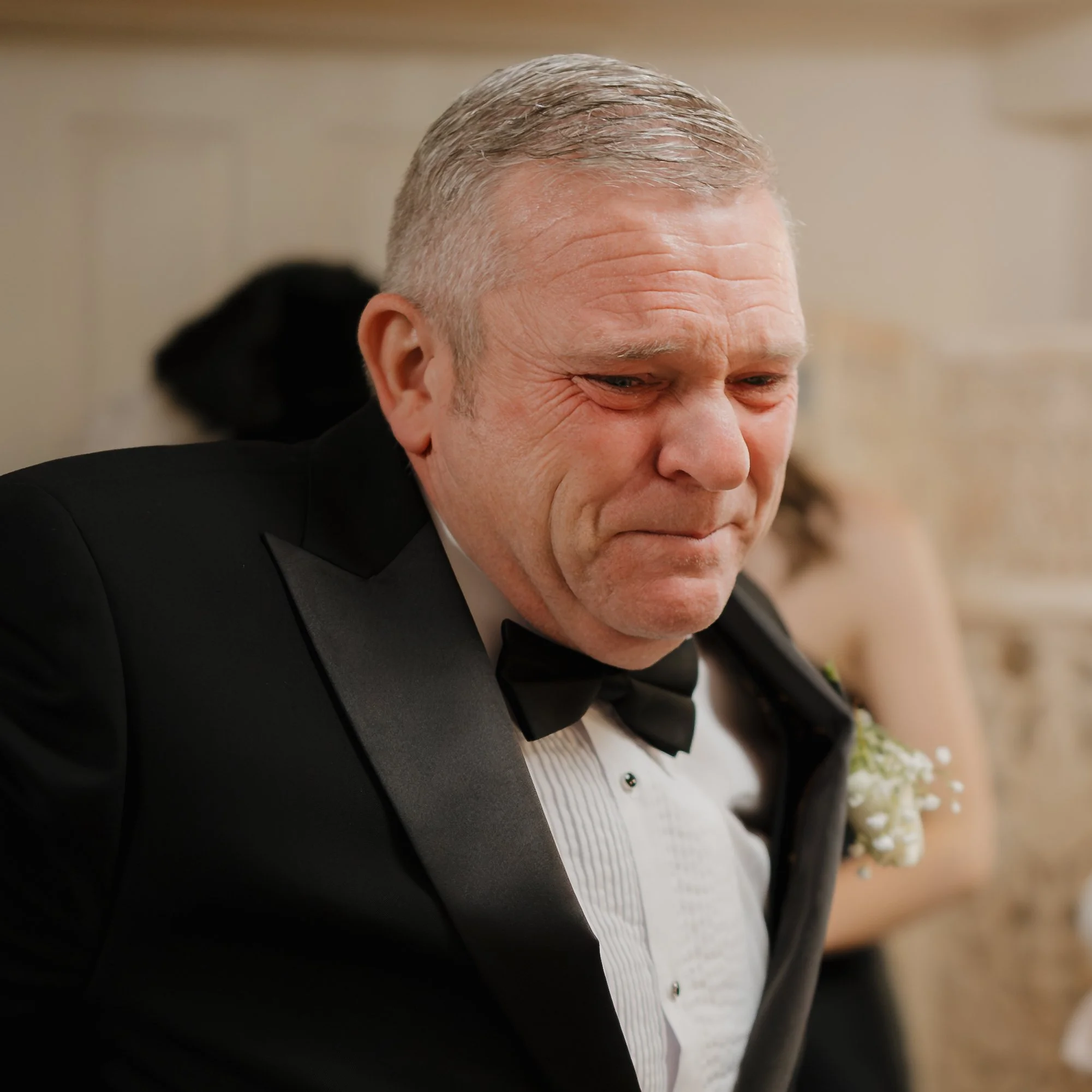 A middle-aged man in a tuxedo, with a black bow tie and a white shirt, sitting in a room with a beige wall. He has a contemplative or emotional expression.