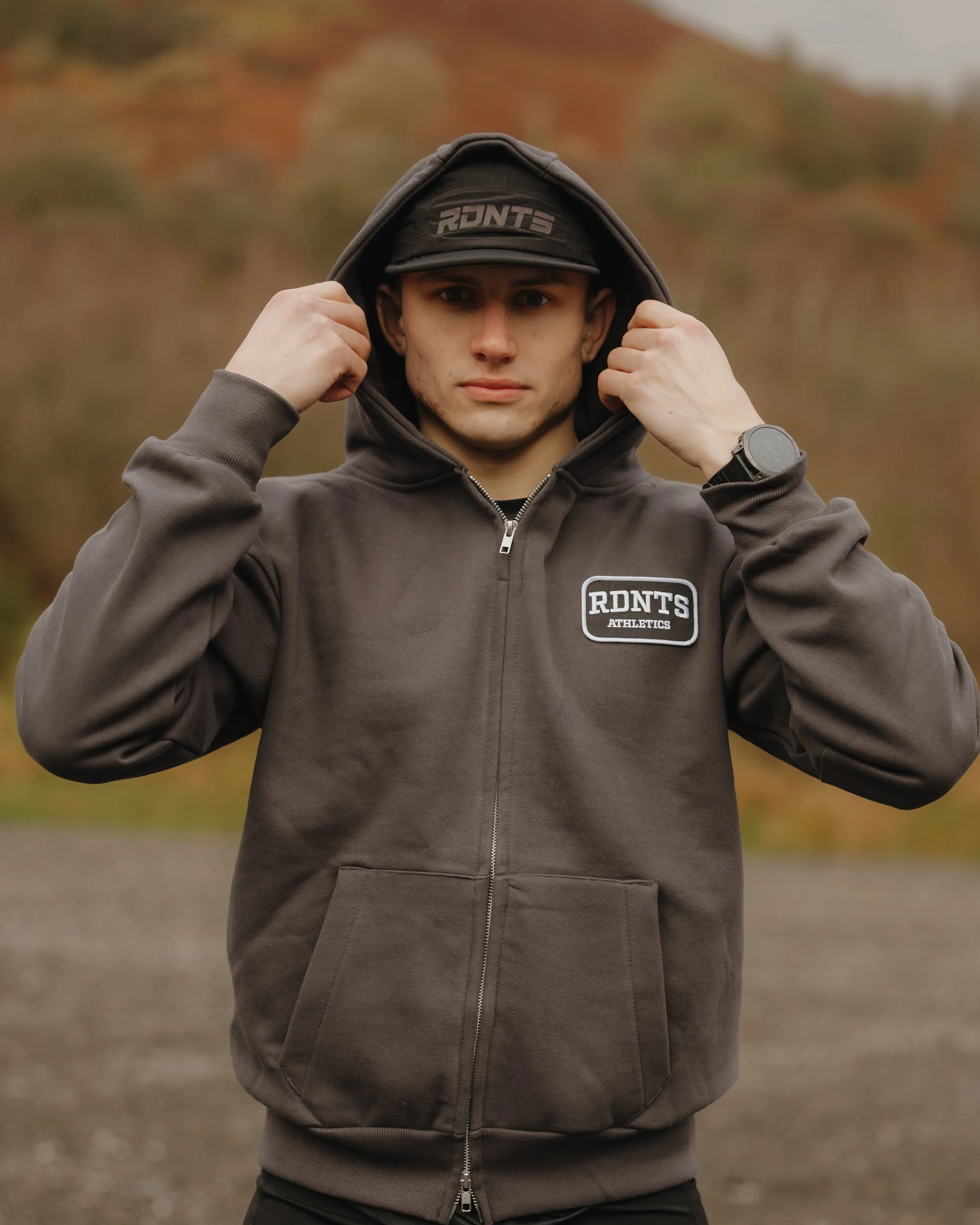 Young man wearing a dark hoodie and baseball cap outdoors, adjusting the hoodie, with autumn-colored trees in the background.