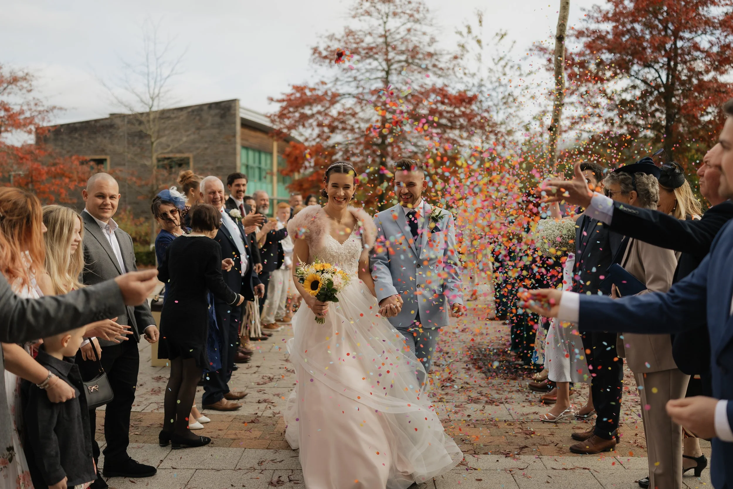 Bride and groom walking hand in hand through a celebration surrounded by guests and colorful confetti, outdoors during autumn.