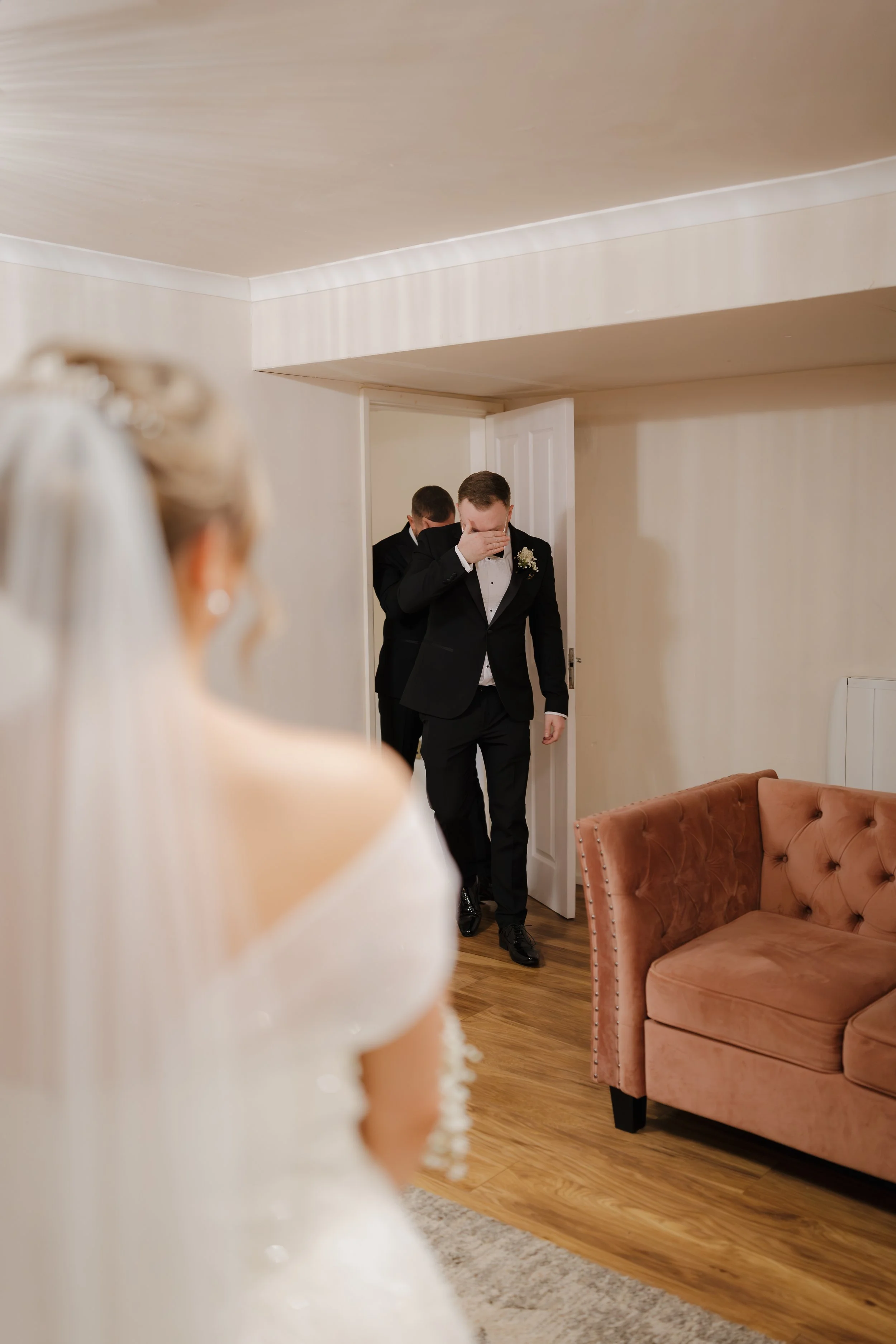 A bride in a white wedding dress with a veil, watching two men in tuxedos, one of whom is covering his face with his hand, as they enter the room.