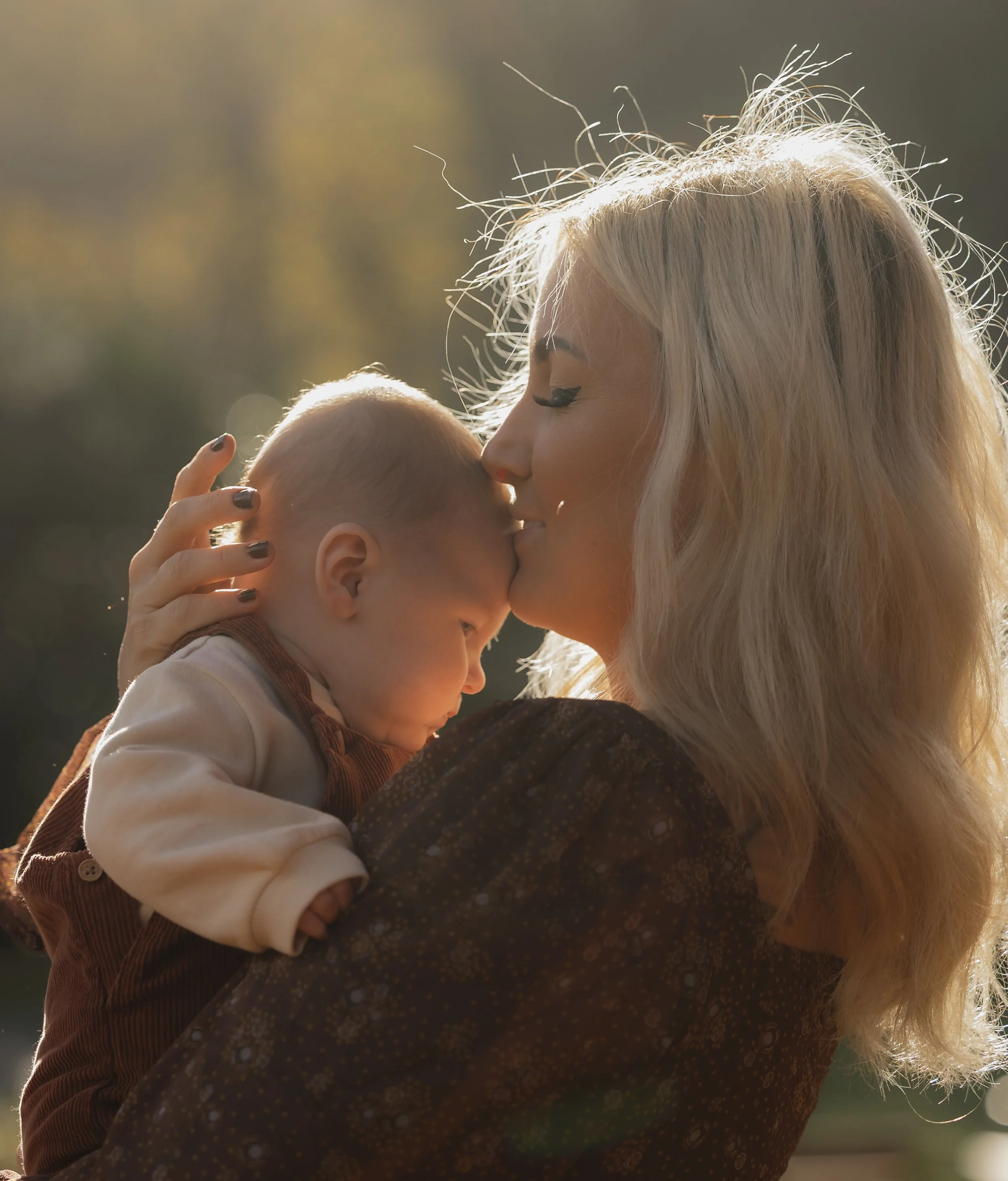 A woman with blonde hair holding a baby close, with the woman kissing the baby's forehead outdoors during sunset.