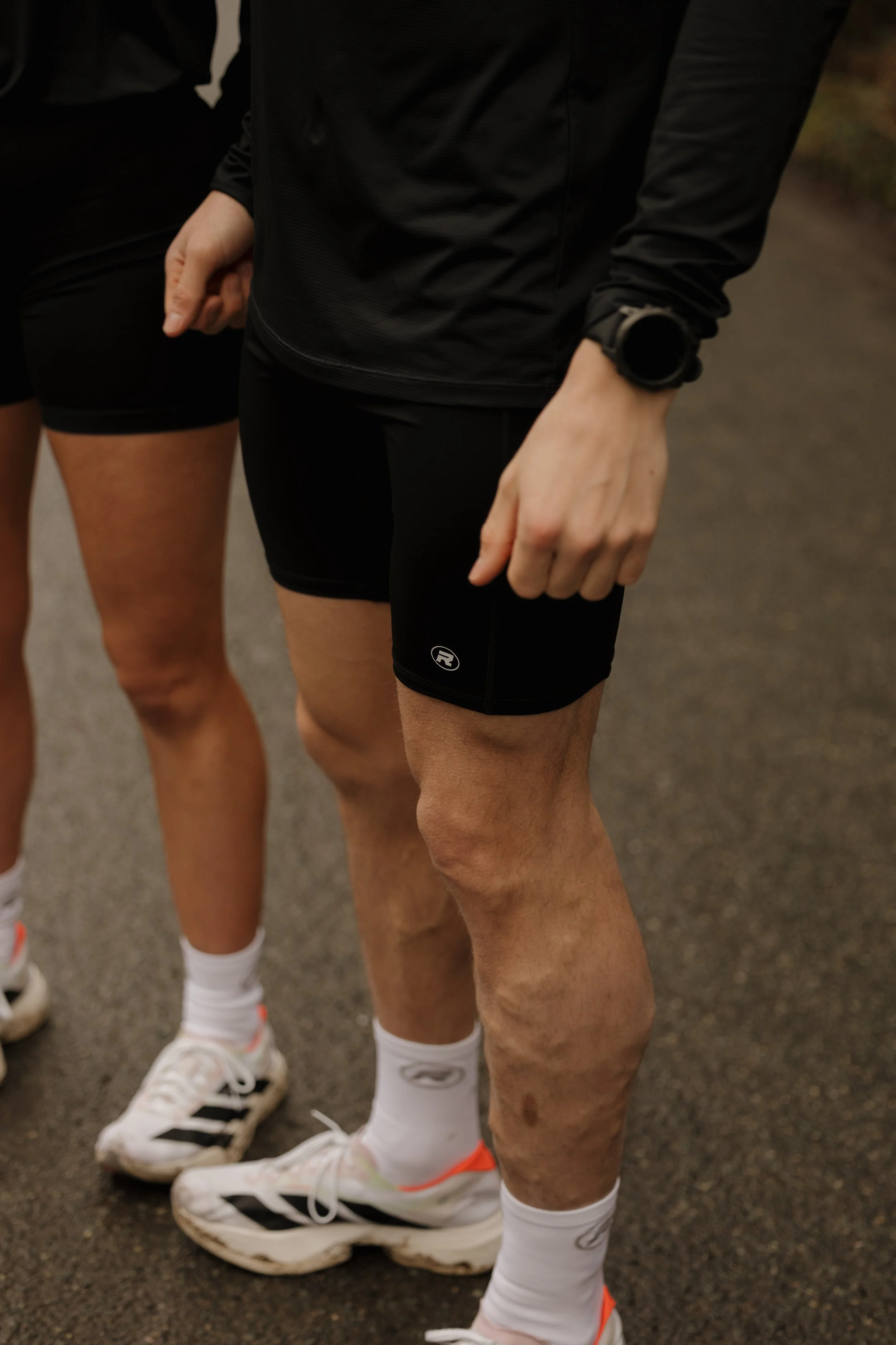 Close-up of two runners in athletic gear, focusing on their legs, shoes, and lower bodies, standing outdoors on a paved path.