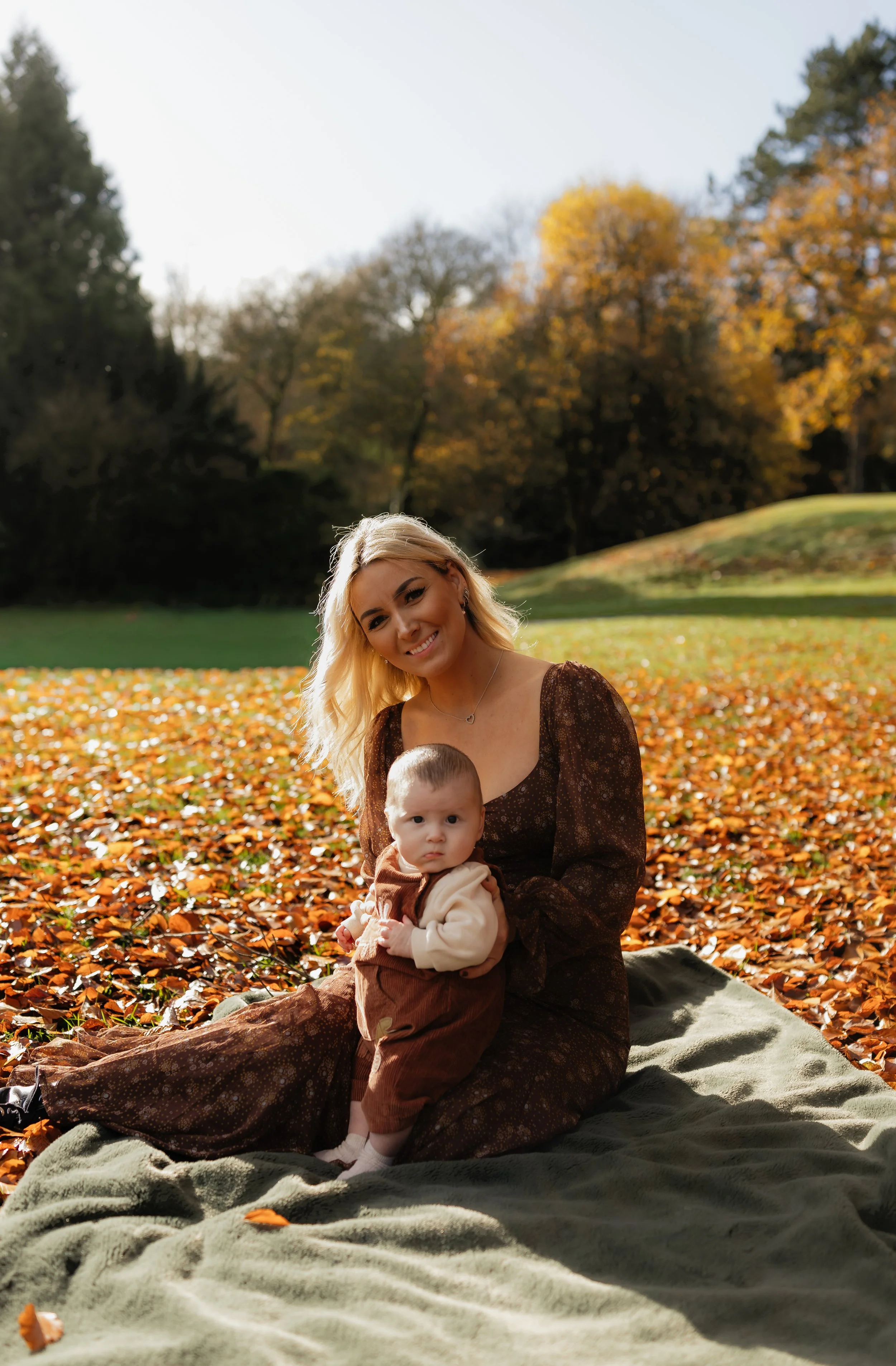 A woman and a young child sitting on a blanket in a park during autumn, surrounded by fallen leaves and trees with colorful foliage.