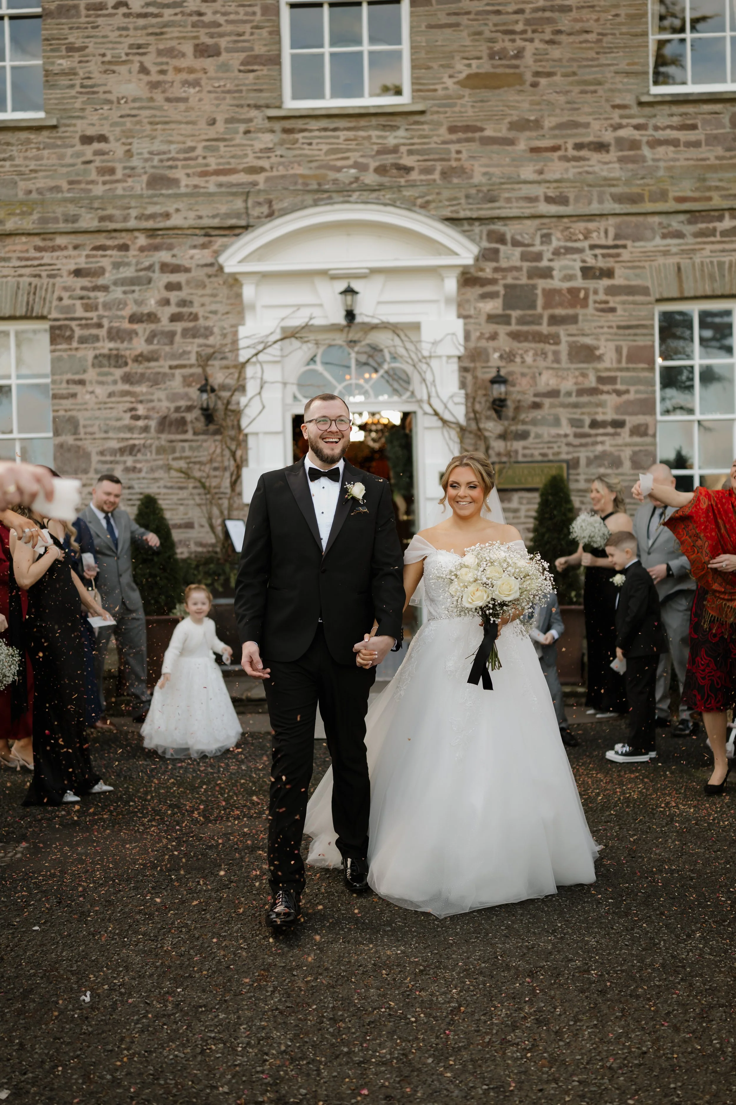 A newlywed couple is walking out of a stone building, holding hands and smiling. The bride is wearing a white wedding dress and holding a bouquet of white roses, while the groom is dressed in a black tuxedo. Guests are celebrating around them, some t