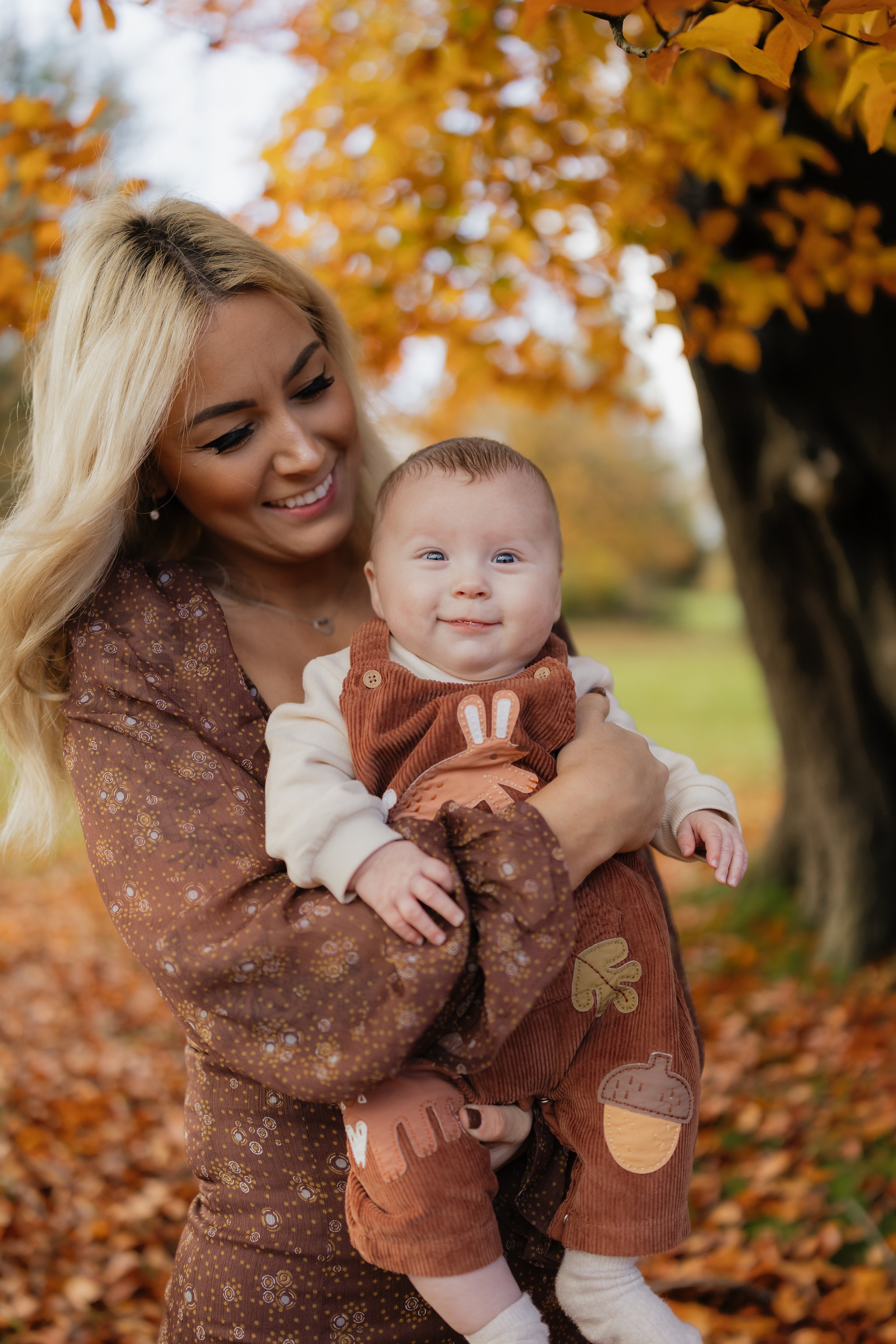 A smiling woman holding a baby outdoors with autumn leaves in the background.