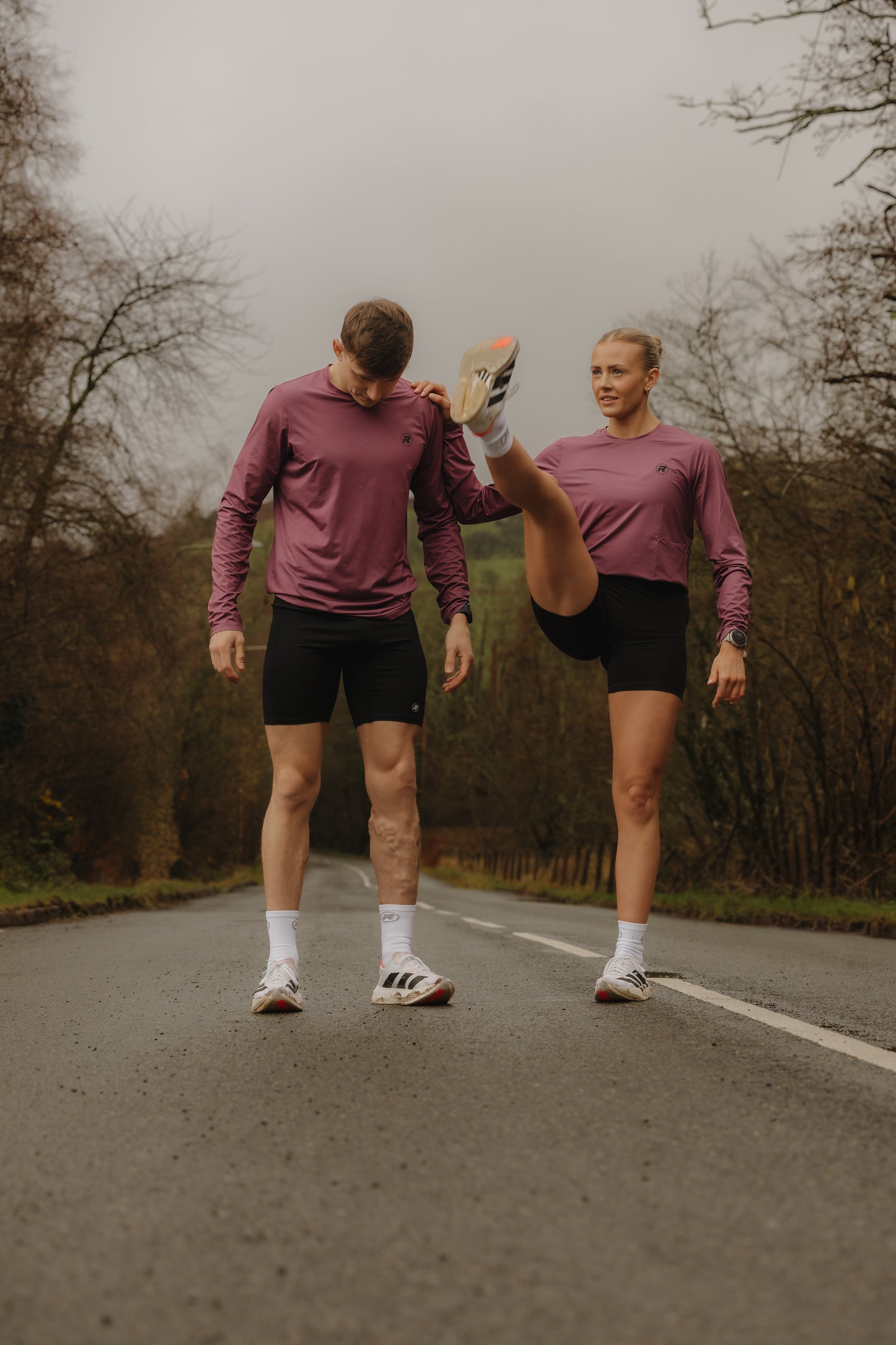 Two people in athletic clothing stretching on a rural road with trees and cloudy sky in the background.